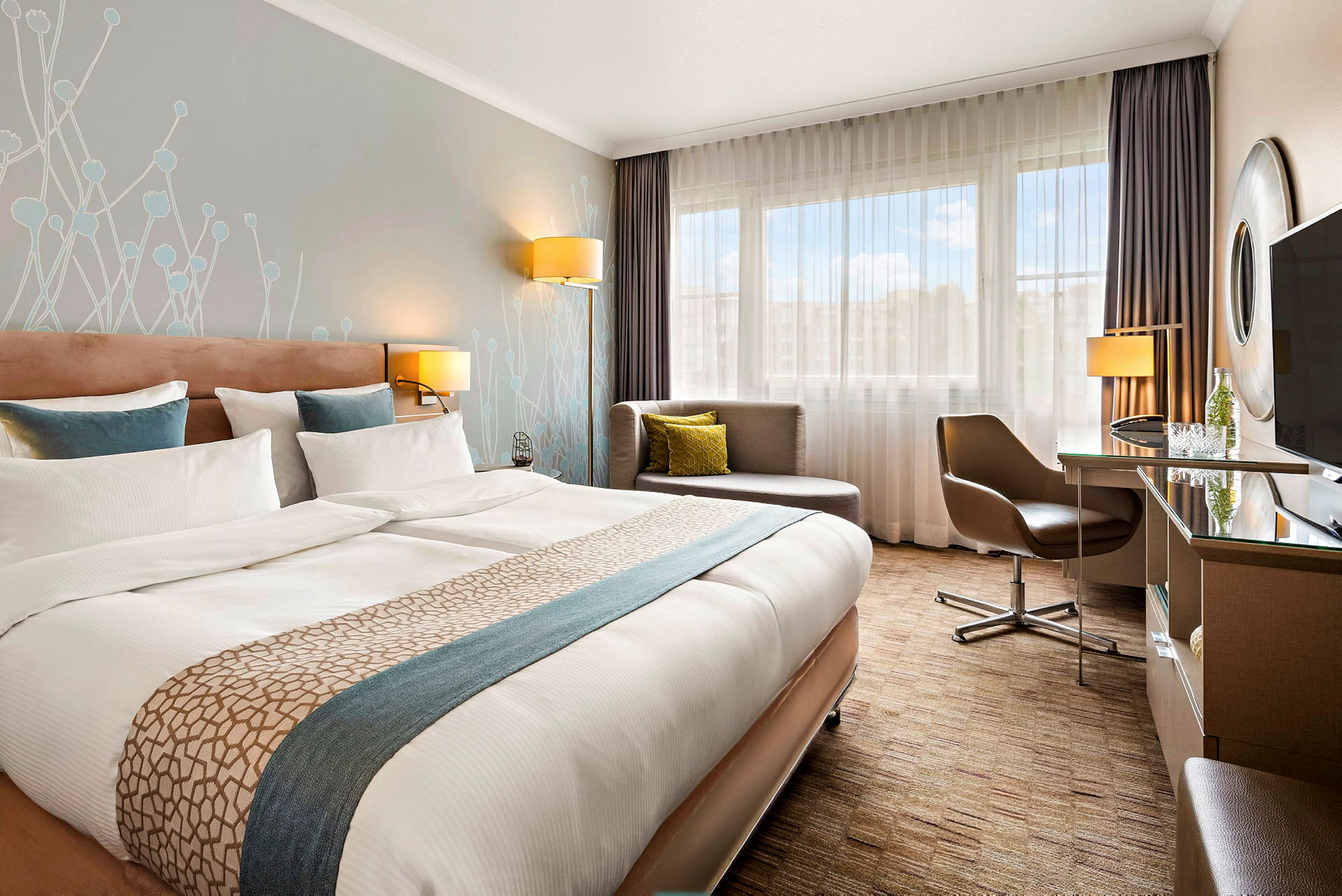 Moderni hotellihuone, jossa suuri parivuode, kirjoituspöytä, tuoli ja istuinryhmä ikkunan vieressä. (Tekoälyn luoma teksti)