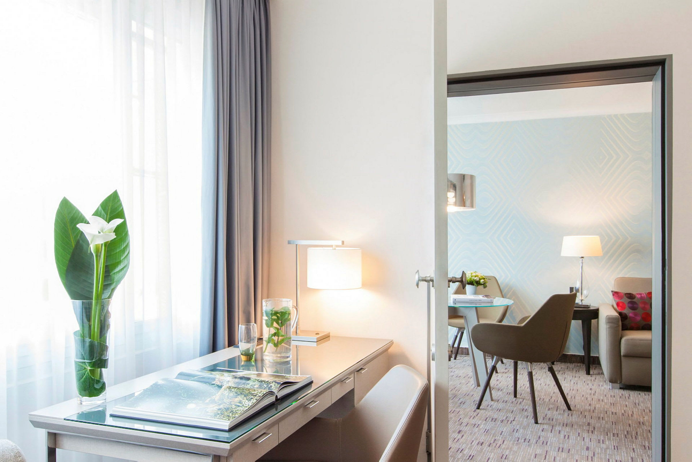 Hotellihuone, jossa pöytä, lamppu ja kukkavaasi sekä istuinryhmä taustalla. (Tekoälyn luoma teksti)