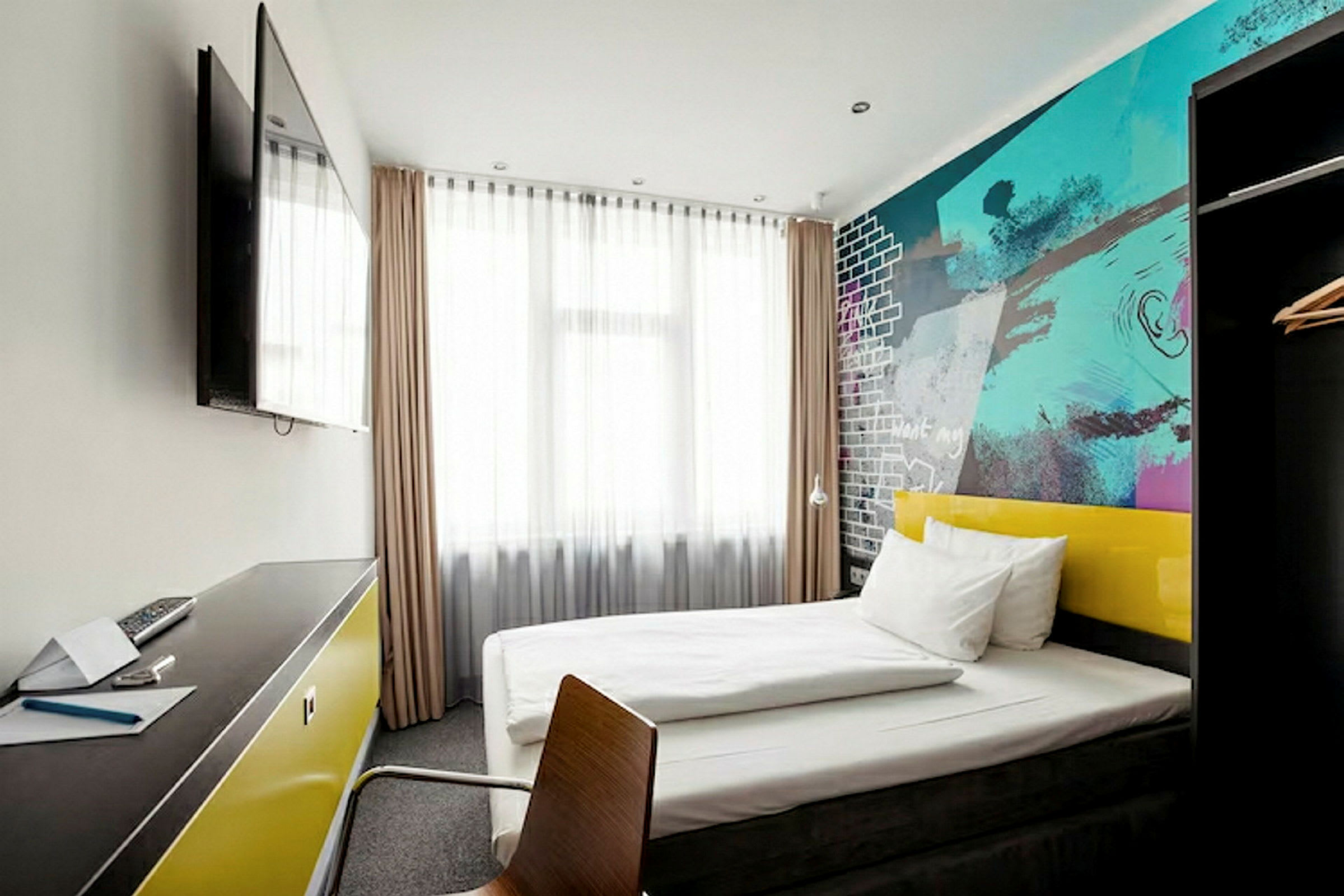 Moderni hotellihuone, jossa on sänky, värikkäät seinät ja työpöytä, jota vastapäätä seinällä on televisio. (Tekoälyn luoma teksti)