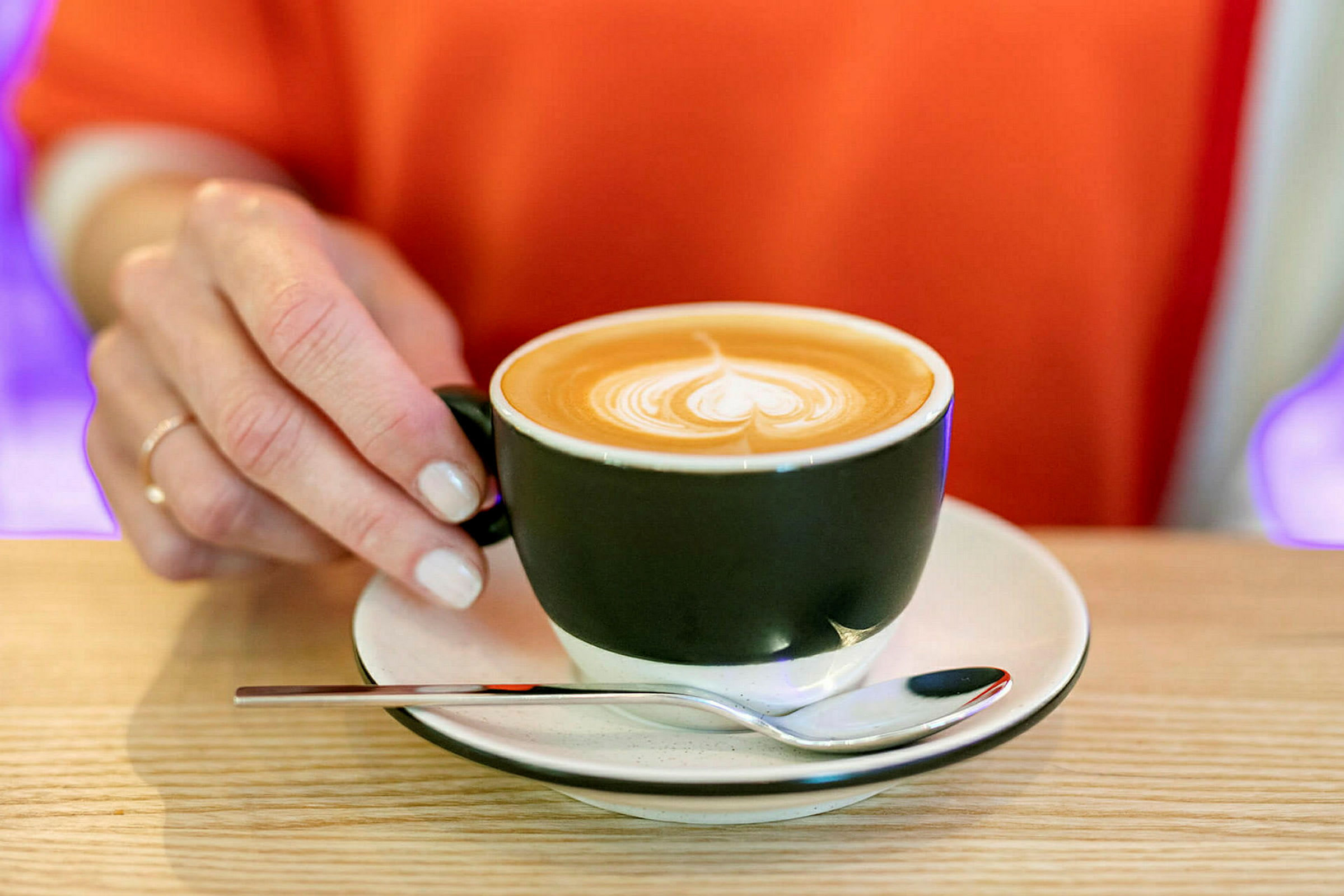 Käsi pitelee mustaa kahvikuppia, jonka alla on lautanen ja lusikka puupöydällä. Cappuccinon pinnalla on latte art. (Tekoälyn luoma teksti)