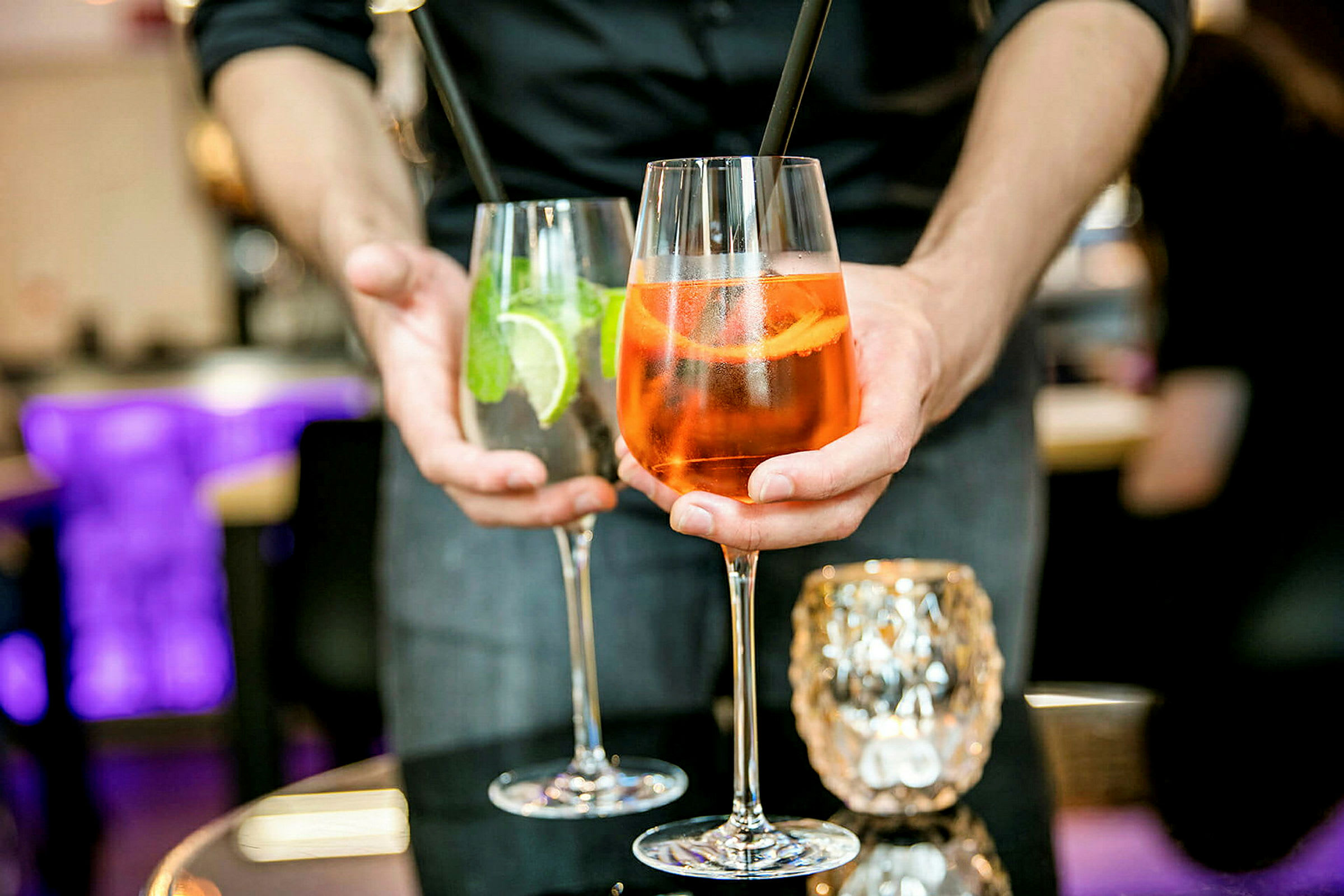 Henkilö pitelee kahta juomaa, joista toisessa lasissa on oranssi cocktail ja toisessa kirkas cocktail limeviipaleiden ja mintun kera. (Tekoälyn luoma teksti)