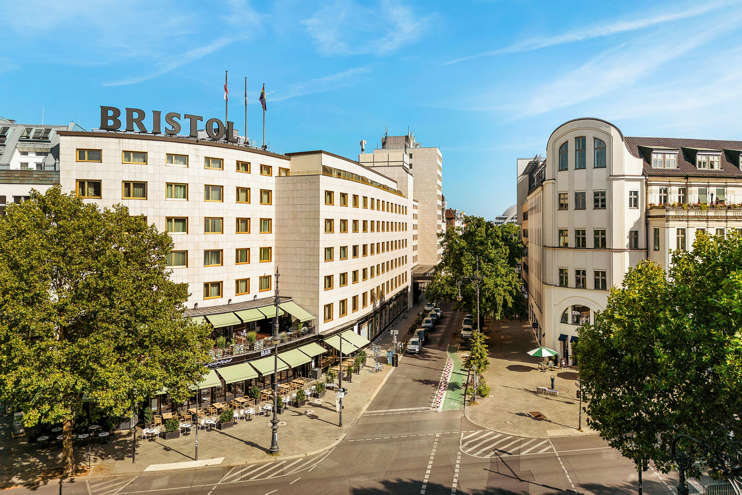 Kaupunkimaisema, jossa on rakennus, jonka katolla lukee "Bristol", ympärillä puita ja katu ulkoilmaravintoloineen. (Tekoälyn luoma teksti)