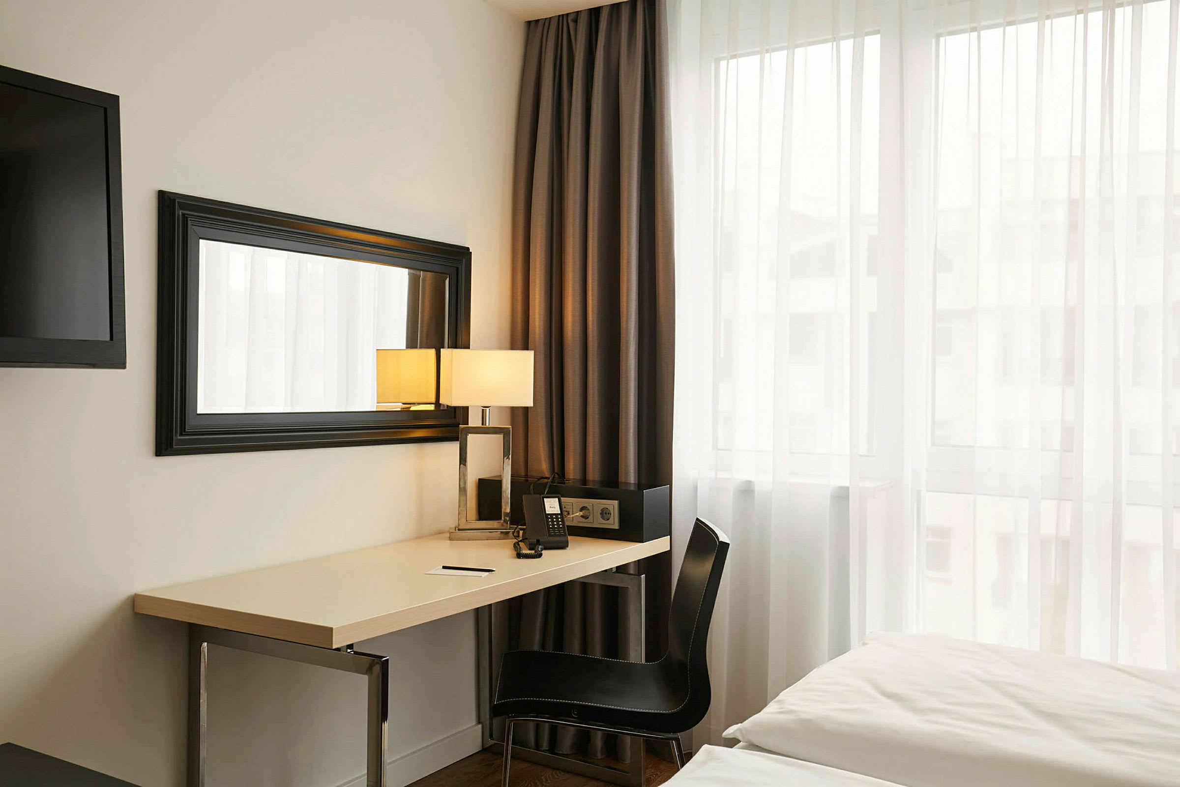 Hotellihuone, jossa on kirjoituspöytä, peili, sänky ja ikkuna. (Tekoälyn luoma teksti)