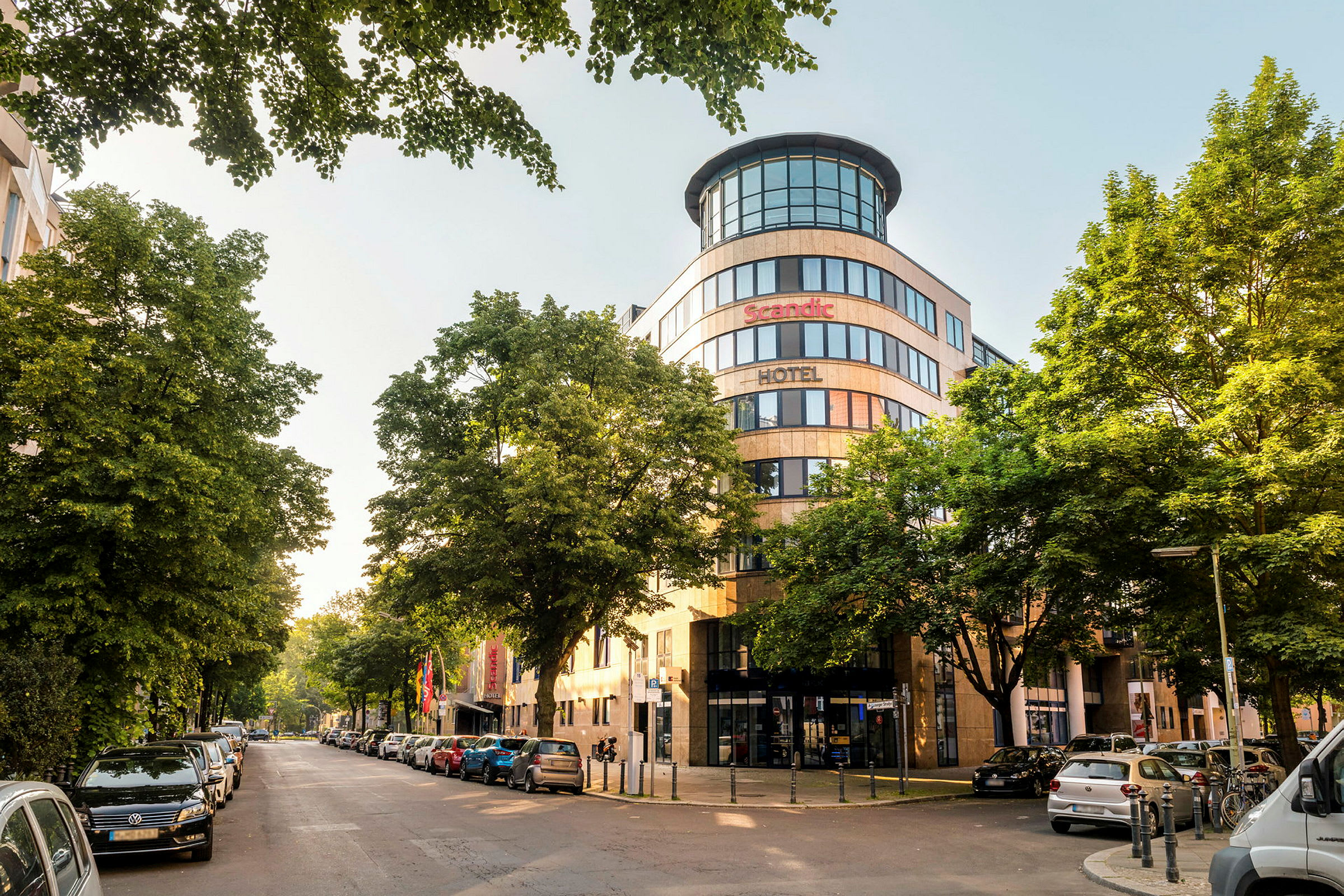 Kaupunginäkymä, jossa on Scandic-hotellin julkisivu, ympärillä puita ja pysäköityjä autoja. (Tekoälyn luoma teksti)