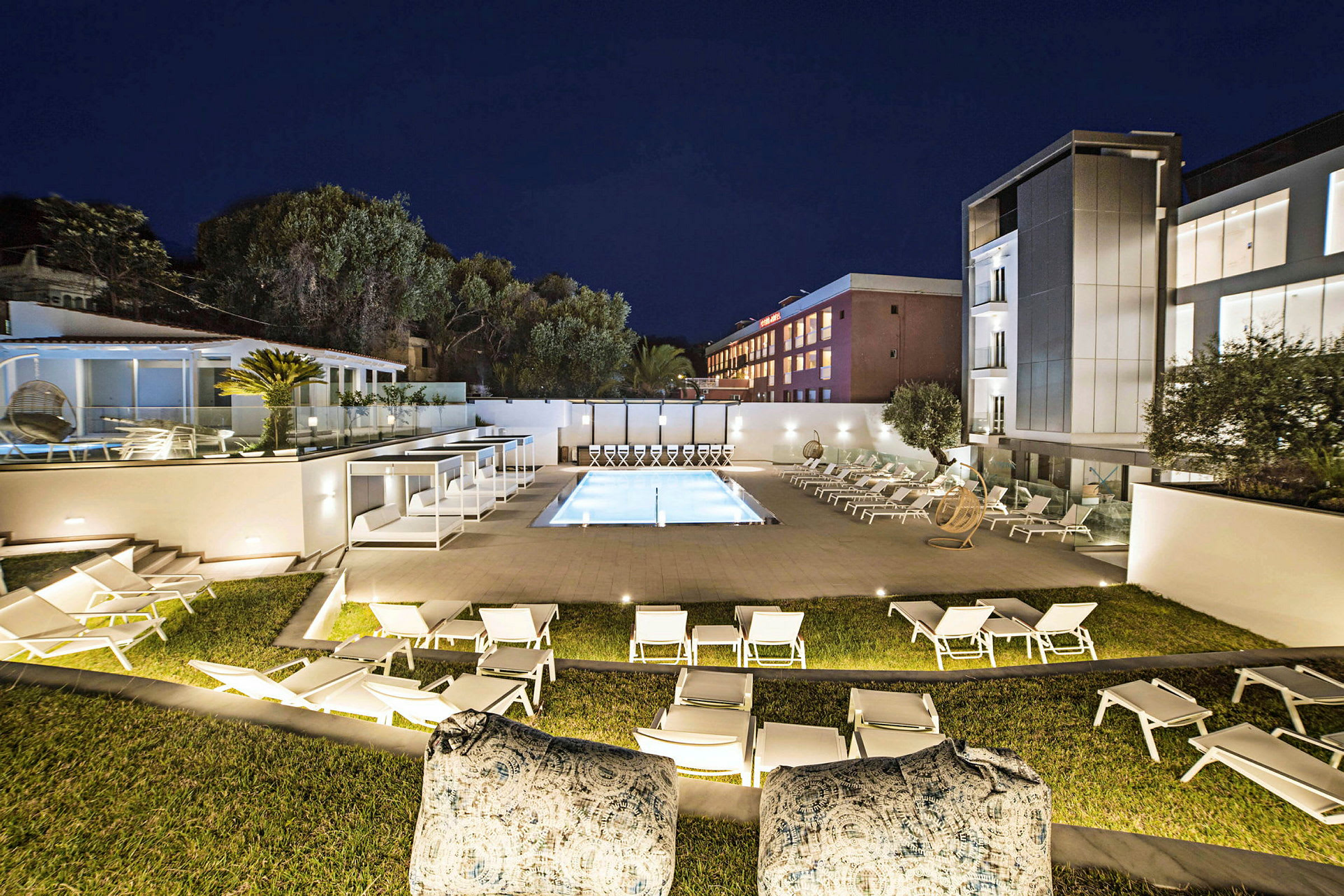 Valaistu hotellialue illalla, jossa on uima-allas, aurinkotuoleja ja moderneja rakennuksia taustalla. (Tekoälyn luoma teksti)