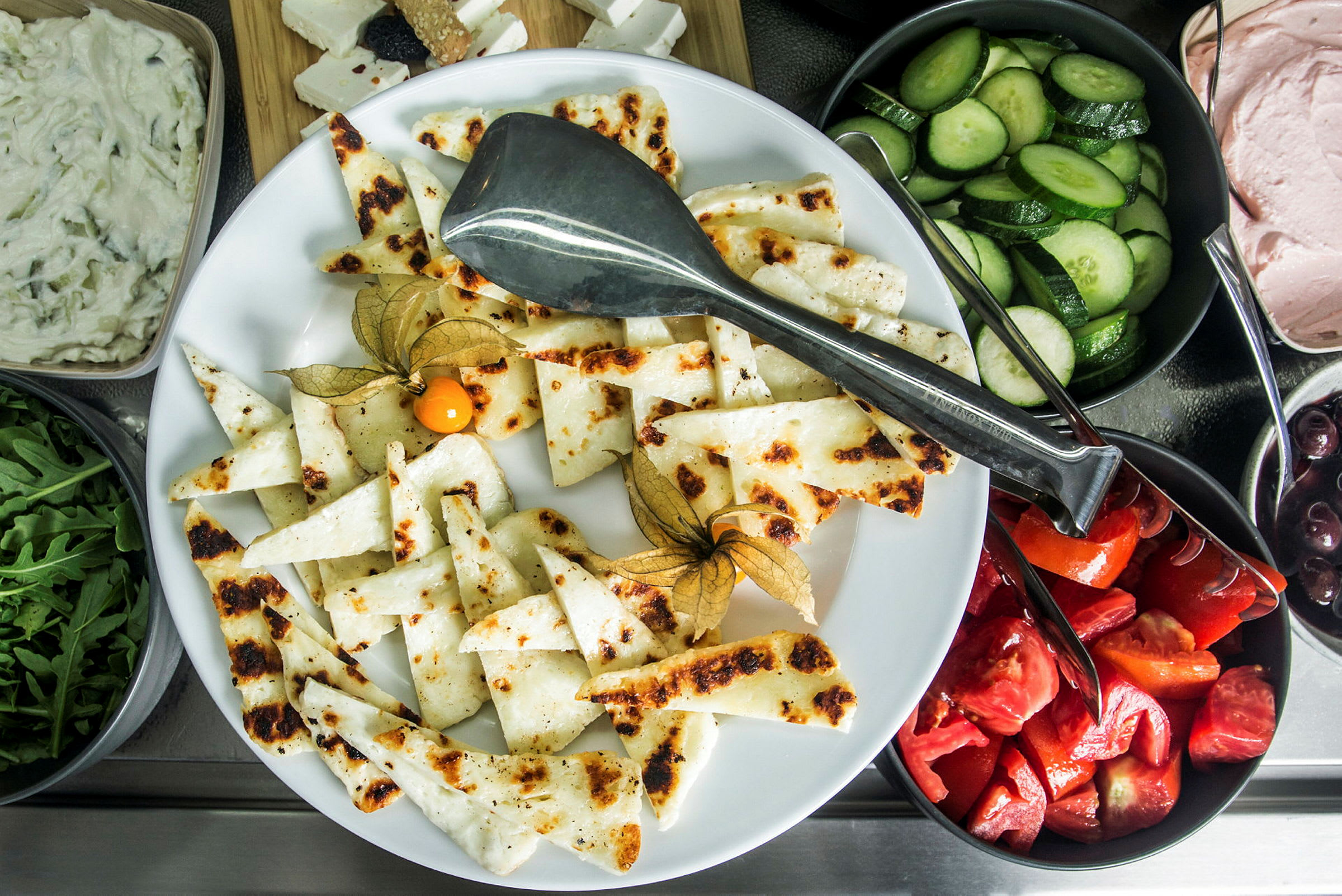Lautanen, jossa grillattua juustoa ja koristeita, ympärillä kulhoja vihanneksia ja kastikkeita. (Tekoälyn luoma teksti)