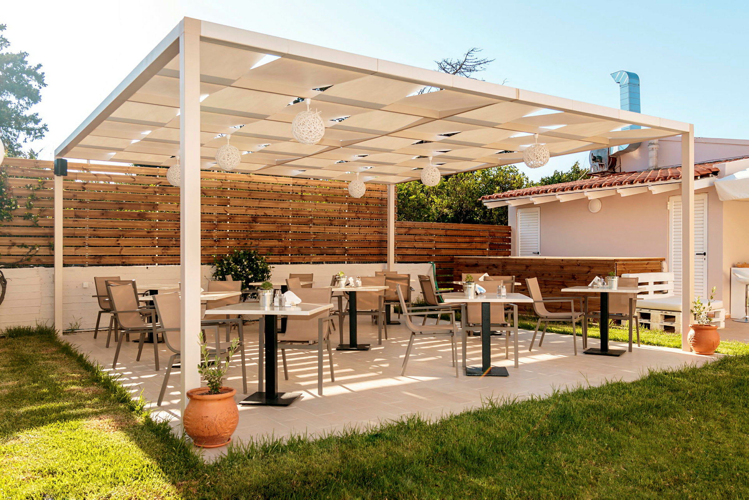 Ulkoterassi, jossa on puurakenteinen pergola, pöytiä ja tuoleja laatoitetulla lattialla, jota ympäröi nurmialue ja aita. (Tekoälyn luoma teksti)