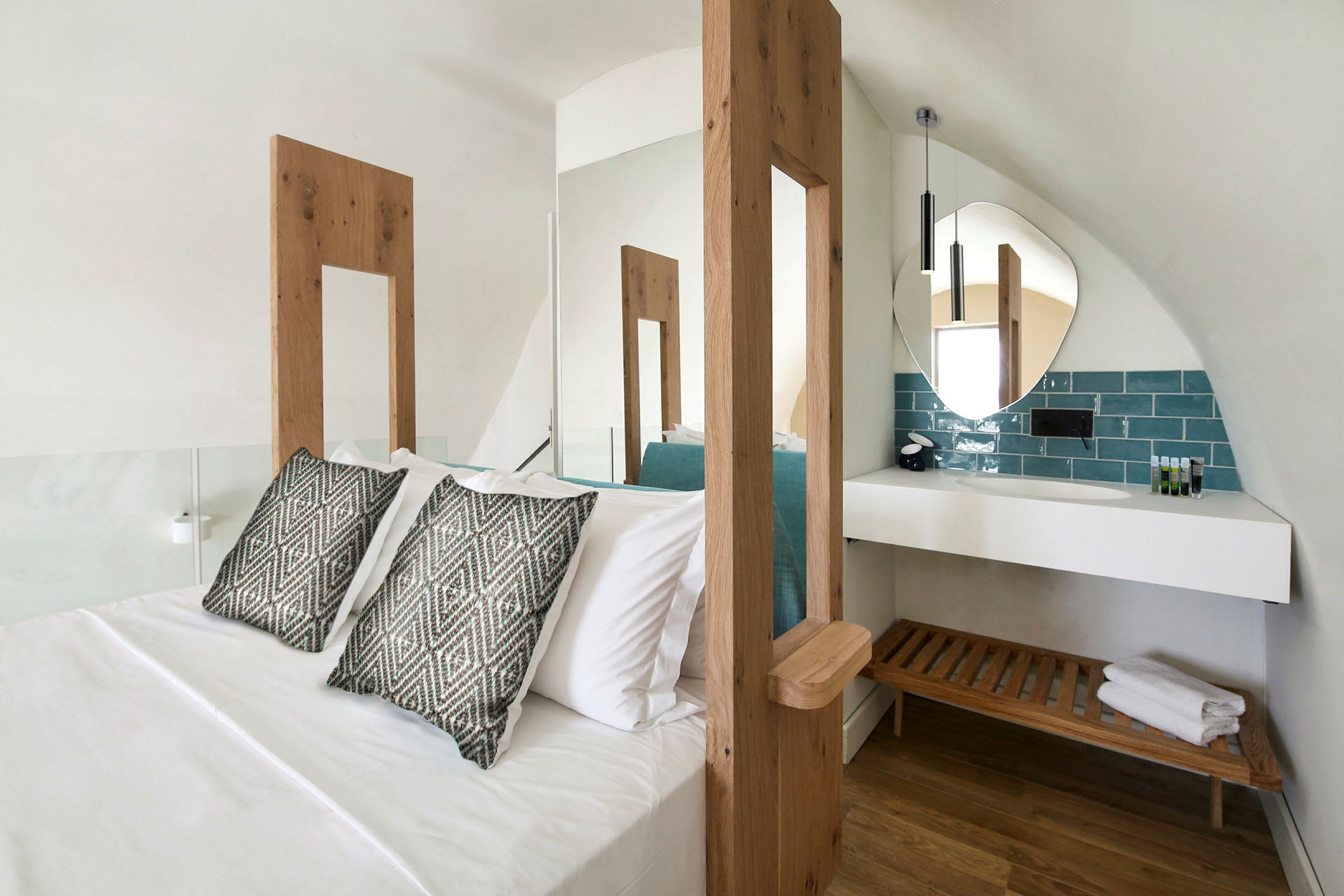 Hotellihuone, jossa suuri sänky, kaksi koristetyynyä ja moderni kylpyhuonealue pyöreällä peilillä ja puisella penkillä valkoisen hyllyn alla. (Tekoälyn luoma teksti)