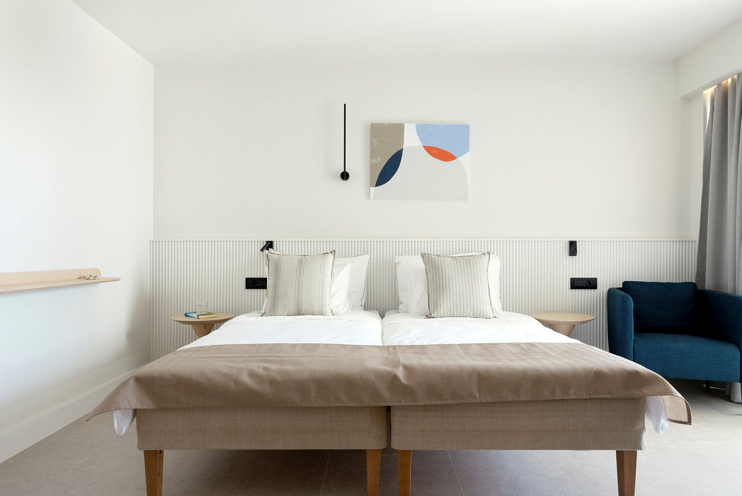 Hotellihuone, jossa on parivuode, sininen nojatuoli ja modernia seinäkoristelua. (Tekoälyn luoma teksti)