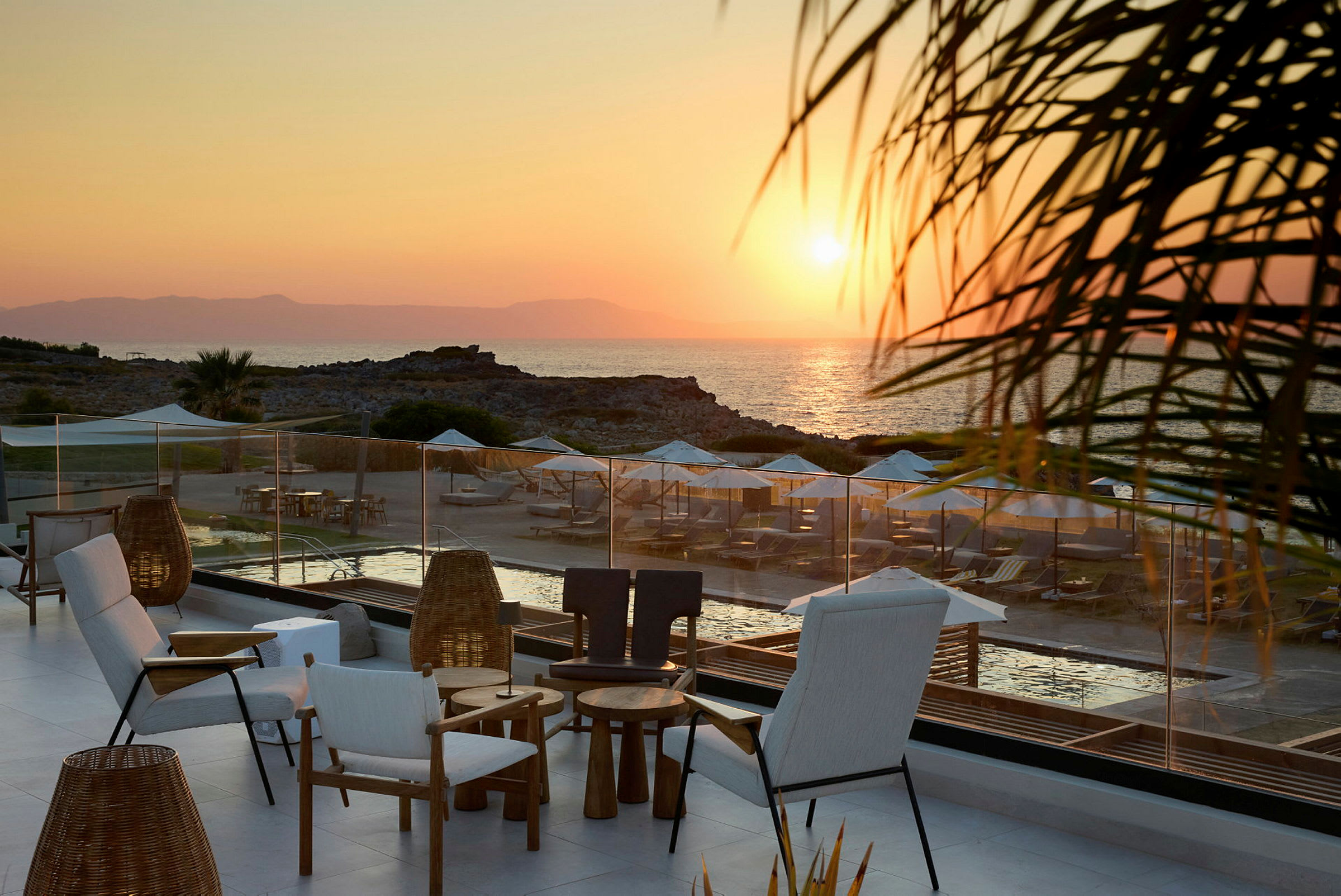 Terassi, jonka etualalla on istumapaikkoja, näkymä rannalle, jossa on aurinkotuoleja ja aurinkovarjoja auringonlaskun aikaan. (Tekoälyn luoma teksti)