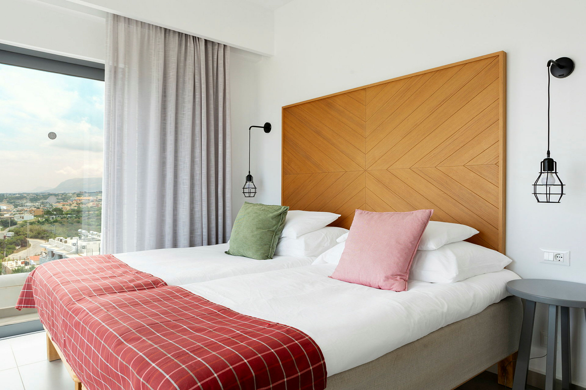 Hotellihuone, jossa kaksi vuodetta, värikkäitä tyynyjä, punaiset peitot ja modernit sisustuselementit, näkymä suurten ikkunoiden läpi. (Tekoälyn luoma teksti)