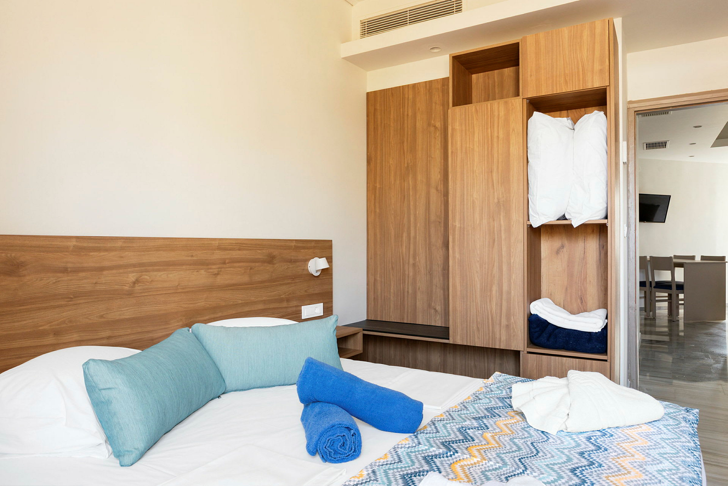 Hotellihuone, jossa sängyssä on sinisiä tyynyjä, sinisiä ja valkoisia pyyhkeitä sekä värikäs kuviollinen päiväpeitto. Taustalla näkyy puunvärinen kaappi ja ovi toiseen huoneeseen. (Tekoälyn luoma teksti)
