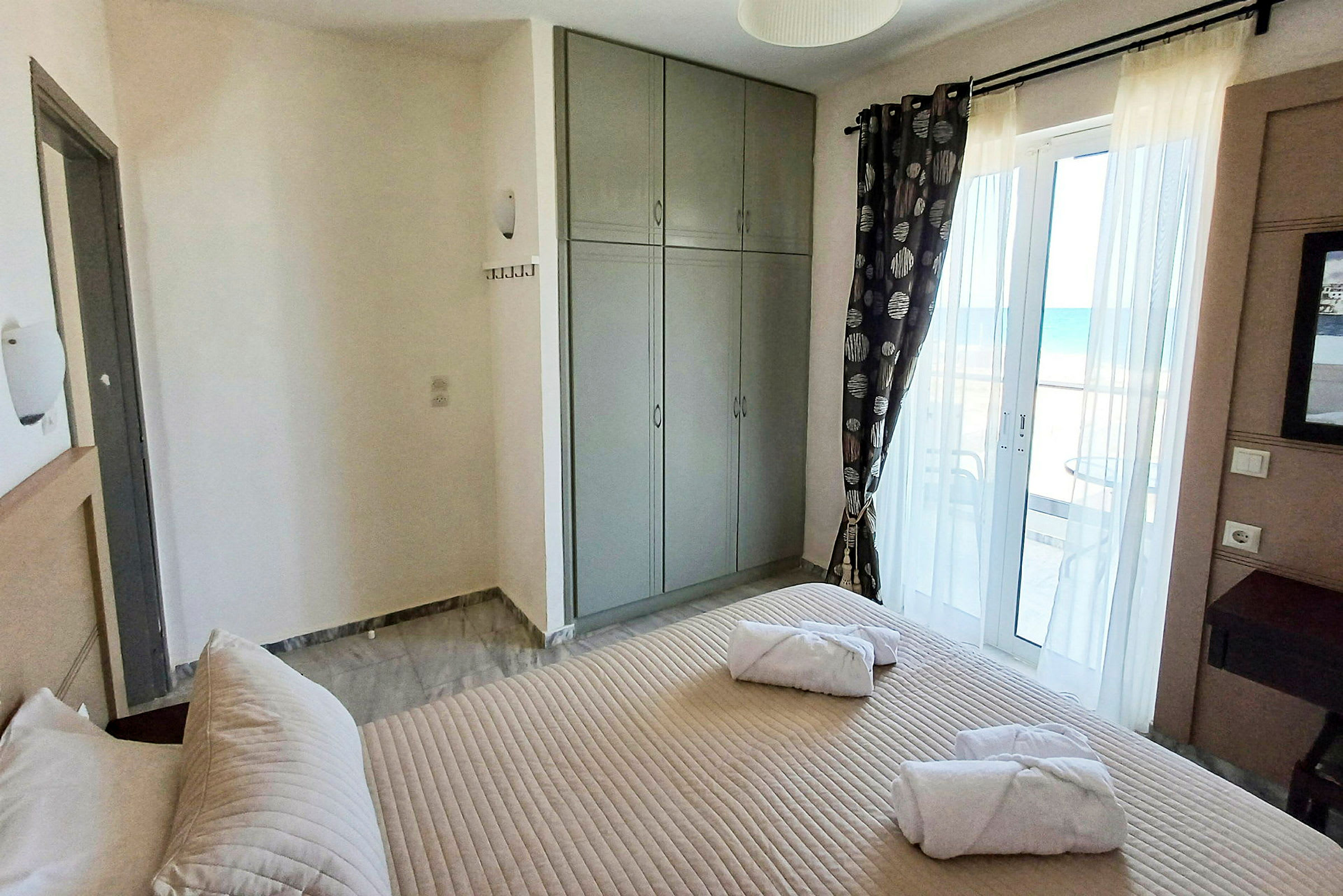 Hotellihuone, jossa sänky, harmaa vaatekaappi ja parvekeovi merinäköalalla. (Tekoälyn luoma teksti)