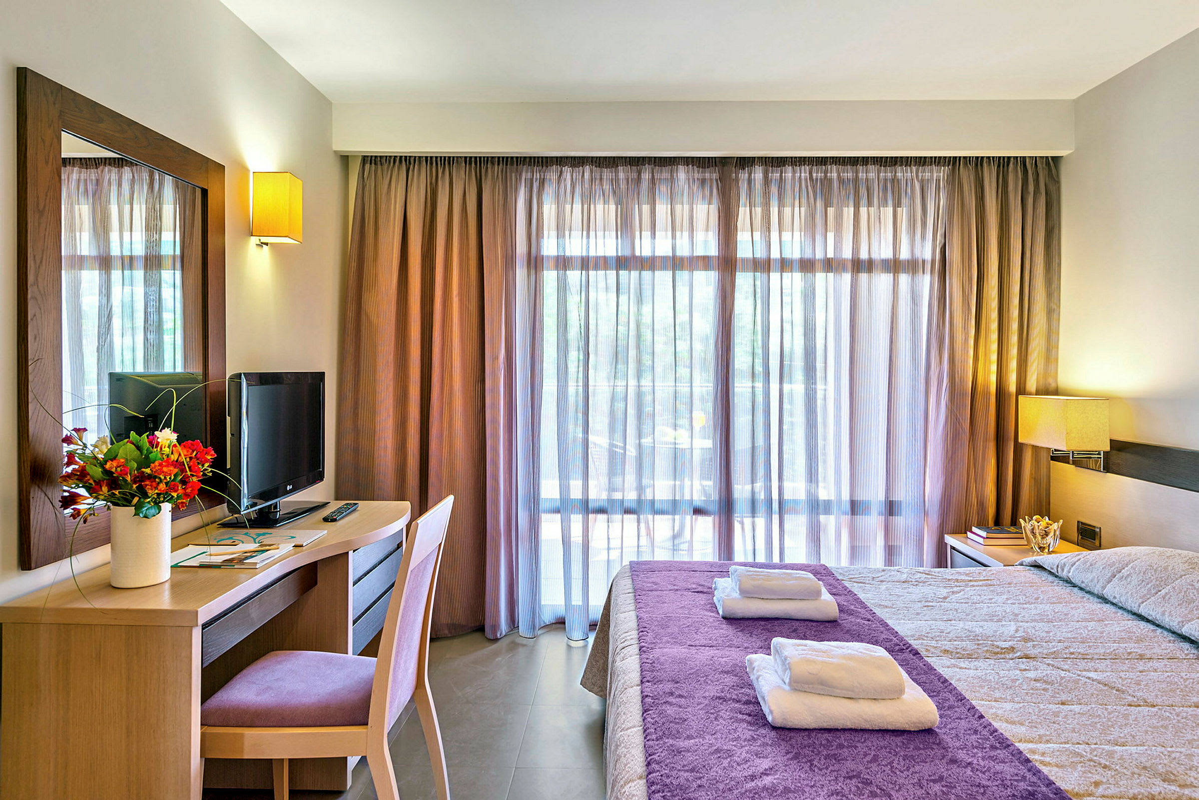 Hotellihuone, jossa sängyllä on violetti päiväpeite, työpöydällä televisio ja kukkakimppu, sekä suuret ikkunat, joissa on läpikuultavat verhot. (Tekoälyn luoma teksti)