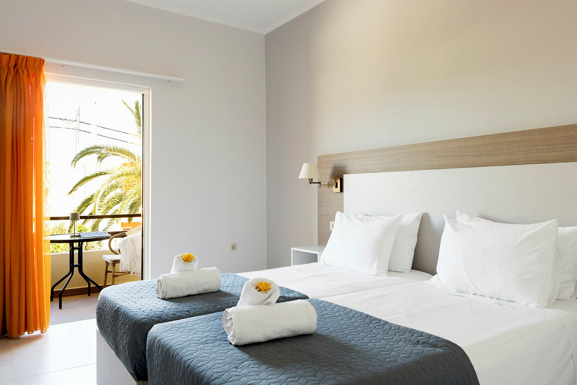 Hotellihuone, jossa kaksi erillistä sänkyä harmailla päiväpeitteillä ja valkoisilla tyynyillä sekä pyyhkeet koristeltuina keltaisilla kukilla. Oransseilla verhoilla varustettu ikkuna avautuu parvekkeelle, josta on näkymä palmuihin. (Tekoälyn luoma teksti)