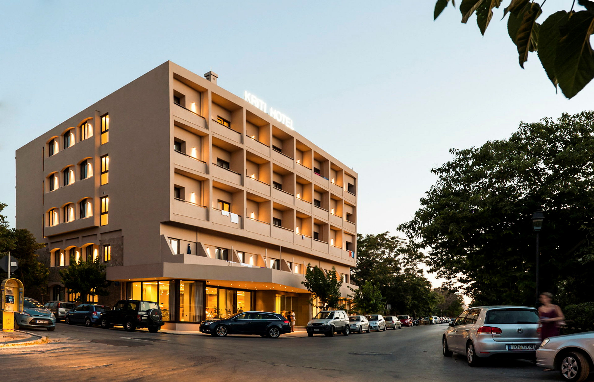 Nelikerroksinen hotellirakennus, valaistu julkisivu, pysäköityjä autoja ympärillä ja taustalla vihreitä puita. (Tekoälyn luoma teksti)