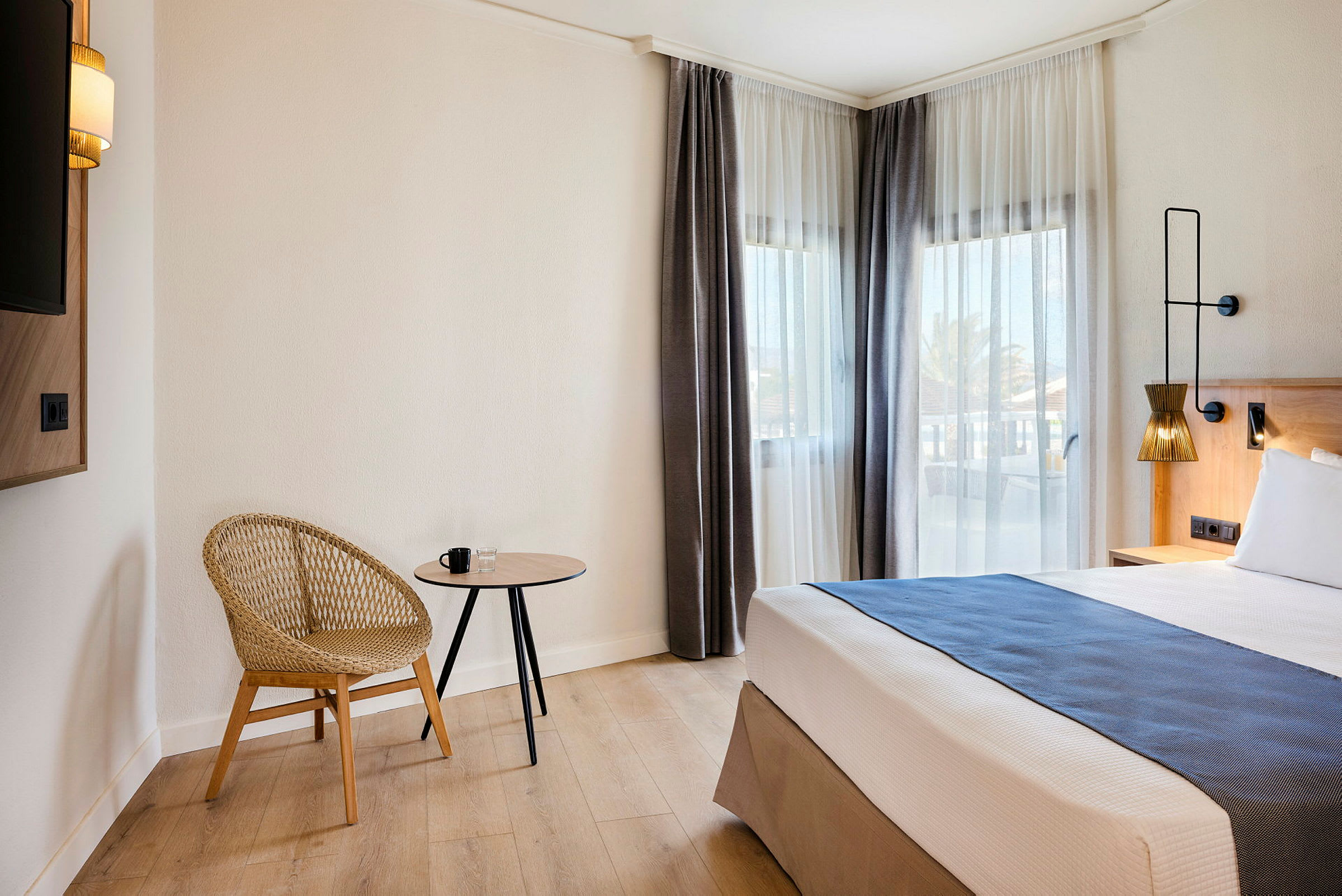 Hotellihuone, jossa moderni tuoli, pieni pöytä ja sänky ikkunoiden vieressä, joissa vaaleat verhot. (Tekoälyn luoma teksti)