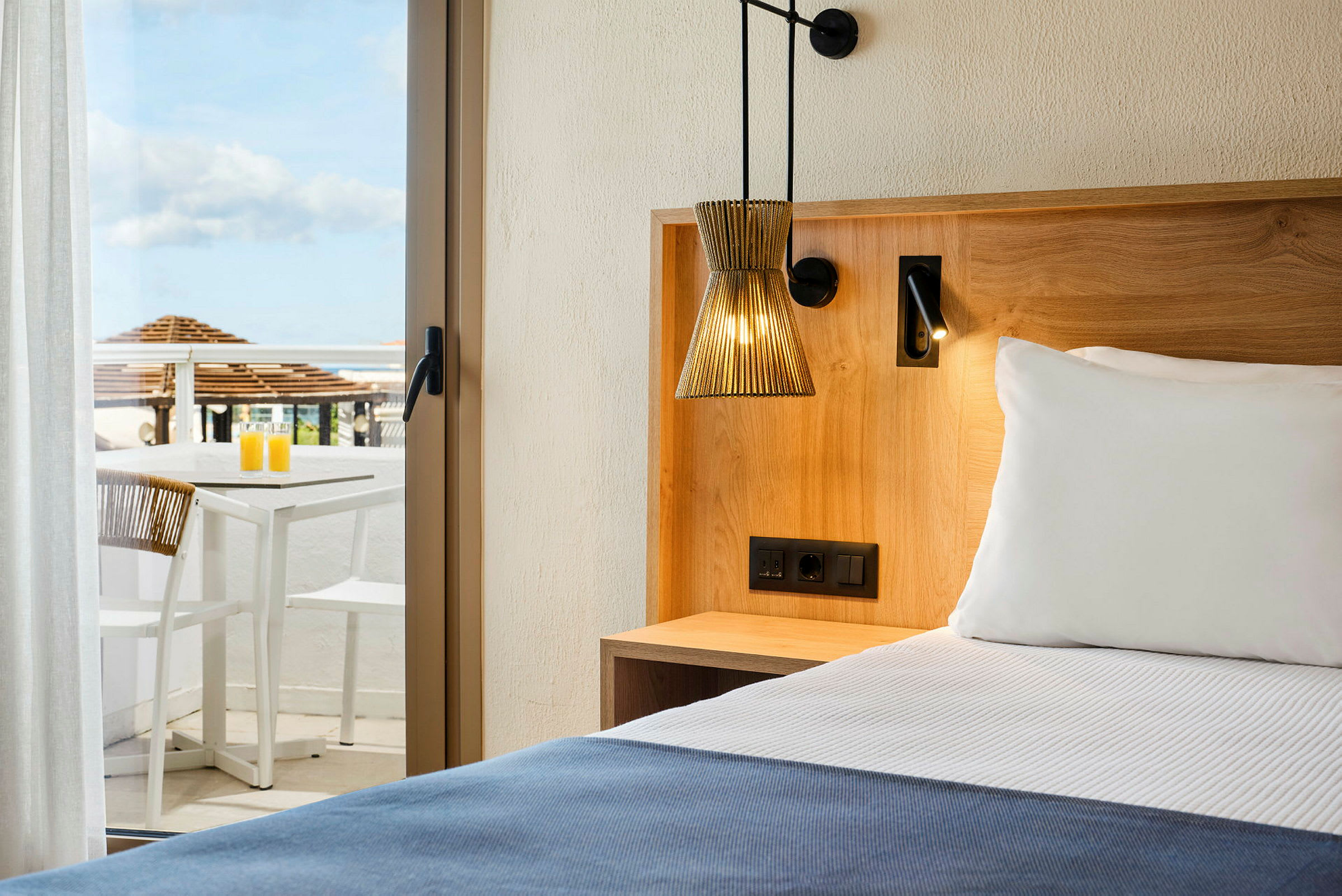 Hotellihuone, jossa on sänky, puupaneelit ja terassi pöydän ja tuolien kanssa taustalla. (Tekoälyn luoma teksti)