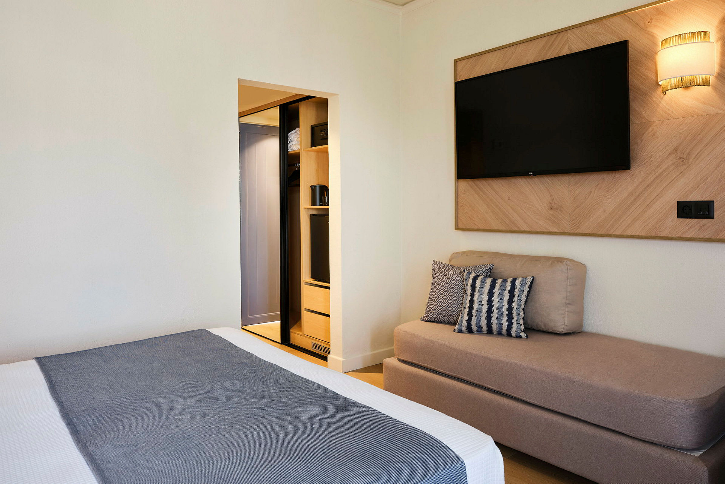 Hotellihuone, jossa on sänky, sohva, seinälle kiinnitetty televisio ja avoin vaatekaappi. (Tekoälyn luoma teksti)