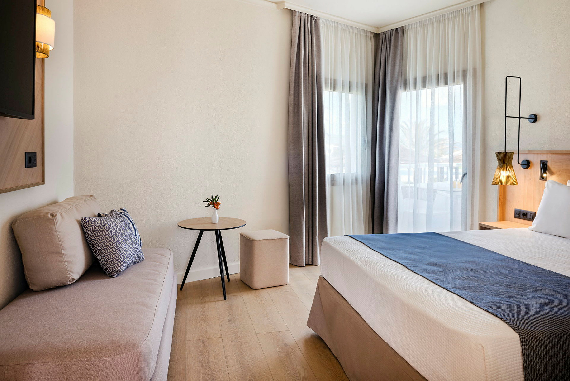 Hotellihuone, jossa on sänky, sohva ja pöytä suurten ikkunoiden vieressä. (Tekoälyn luoma teksti)