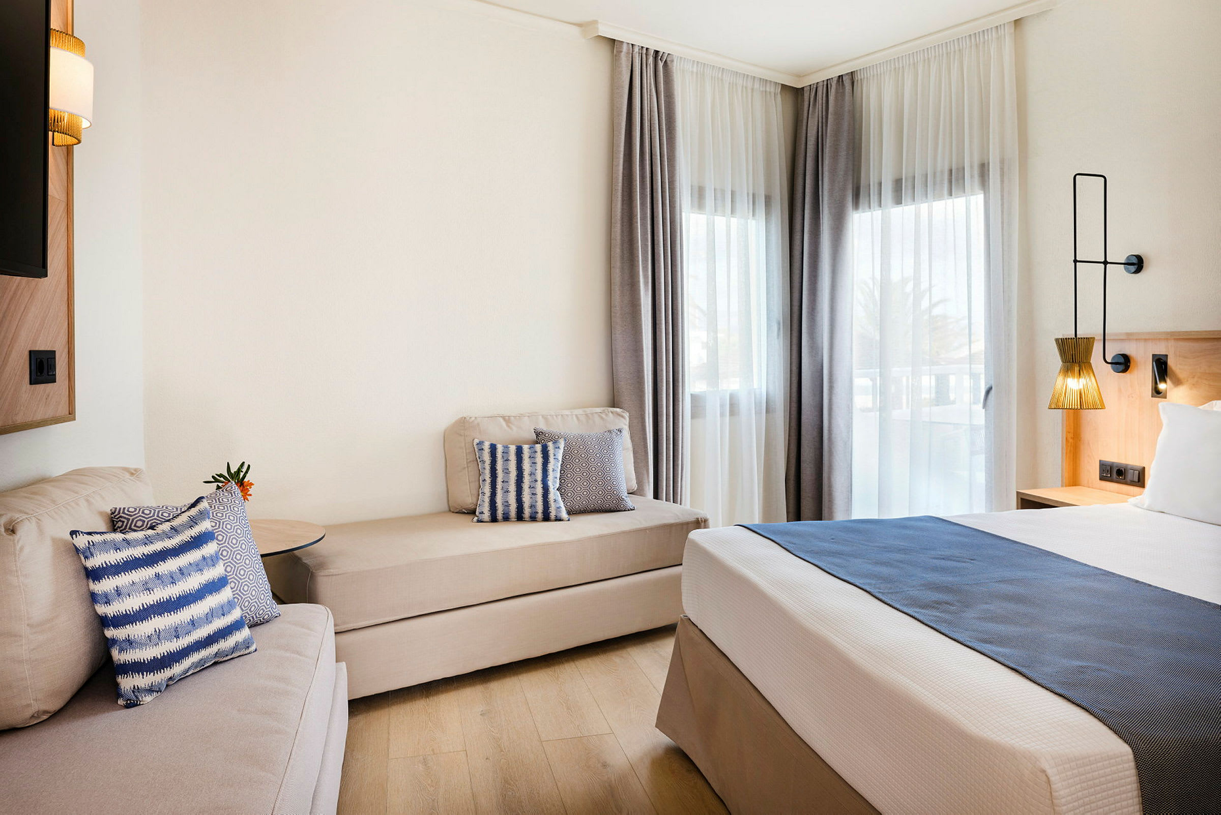 Moderni hotellihuone, jossa on sänky, sohva ja ikkunat vaaleilla verhoilla. (Tekoälyn luoma teksti)