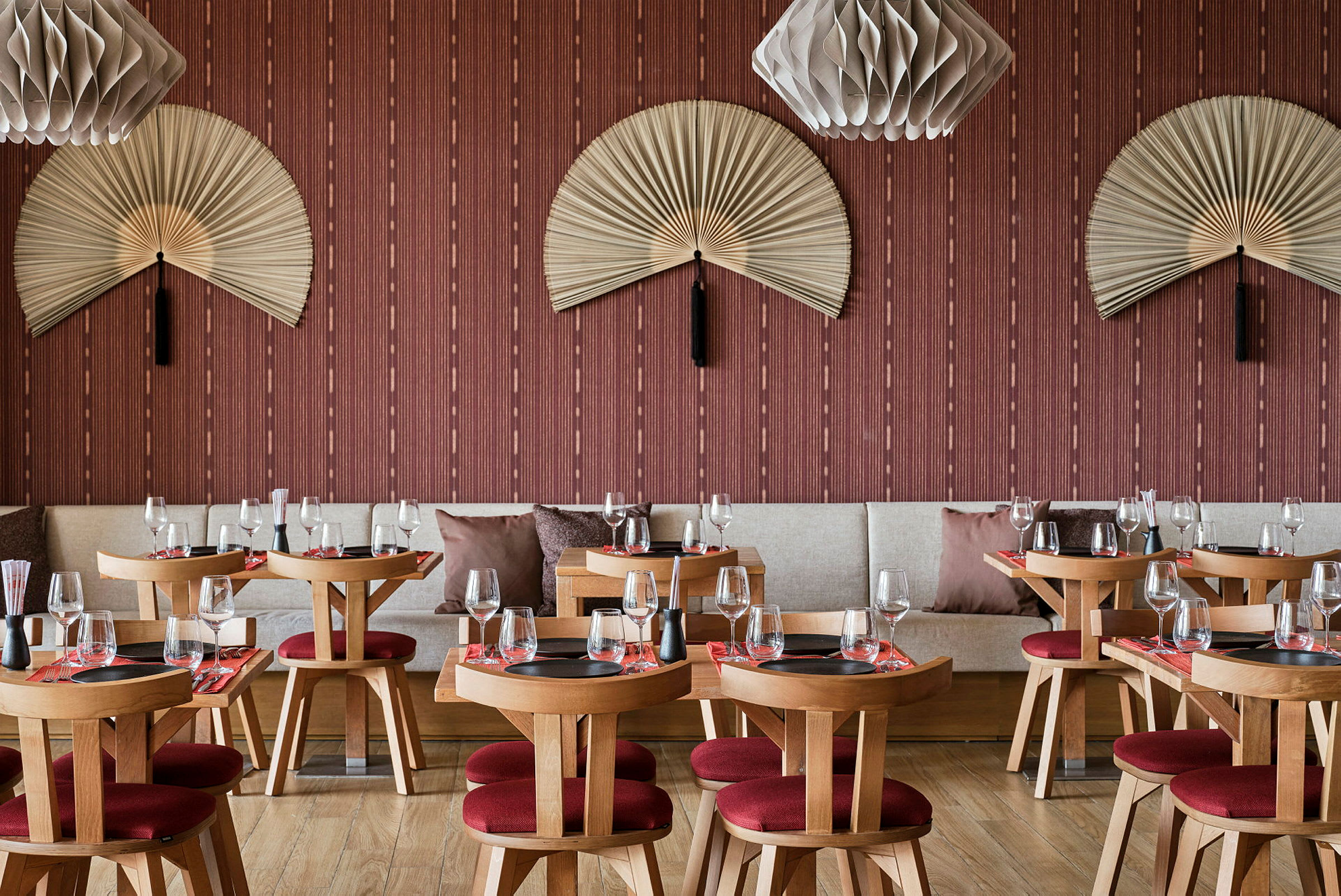 Ravintola, jossa on puupöytiä ja punaisia tuoleja, koristeltu seinälle ripustetuilla viuhkoilla ja moderneilla lampuilla. (Tekoälyn luoma teksti)