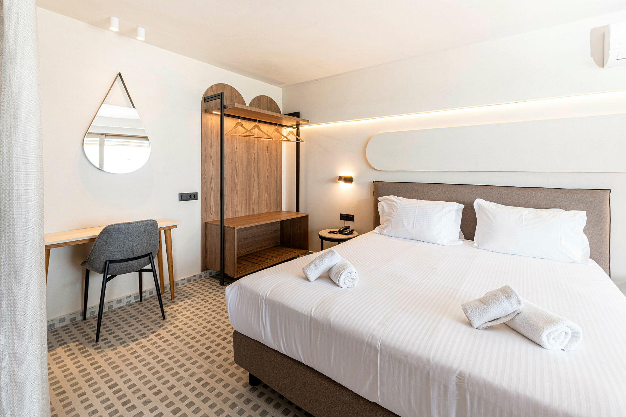 Hotellihuone, jossa on sijattu sänky, kirjoituspöytä tuolilla, seinäpeili ja avoin vaatekaappi. (Tekoälyn luoma teksti)