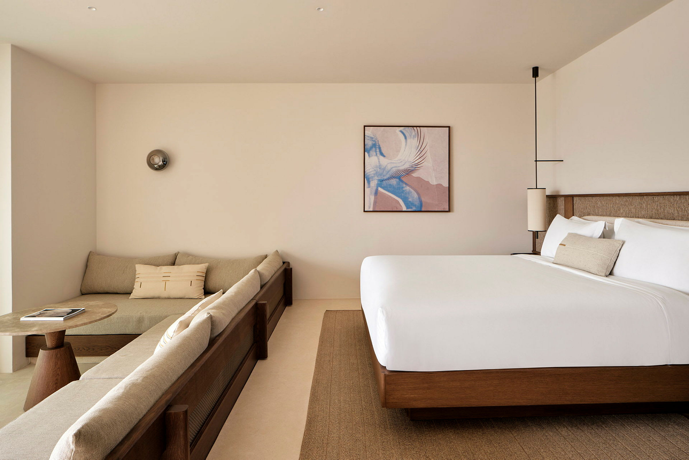Moderni hotellihuone, jossa beige sohva, pyöreä sohvapöytä ja suuri sänky. Näkyvillä on taulu sekä sisustusvalaisimia. (Tekoälyn luoma teksti)
