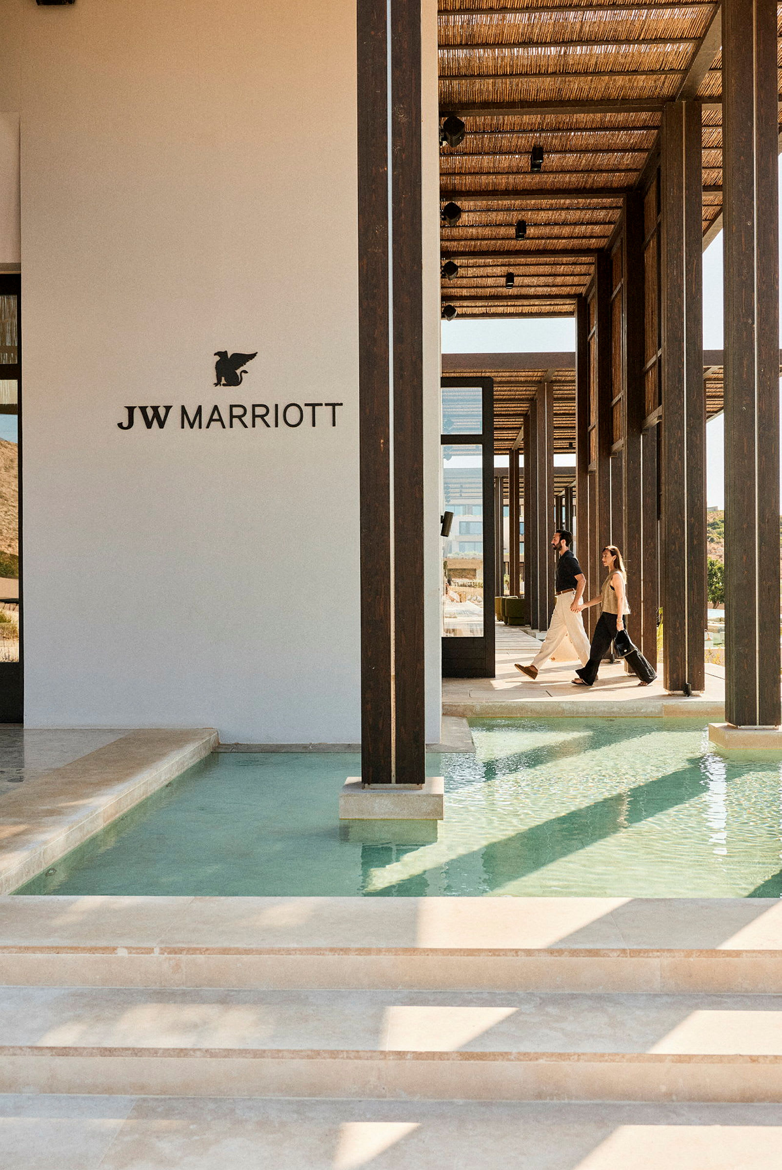 Kuva modernista rakennuksesta, jossa on JW Marriott-logo ja ihmisiä kävelemässä käytävällä, jonka molemmin puolin on vettä. (Tekoälyn luoma teksti)