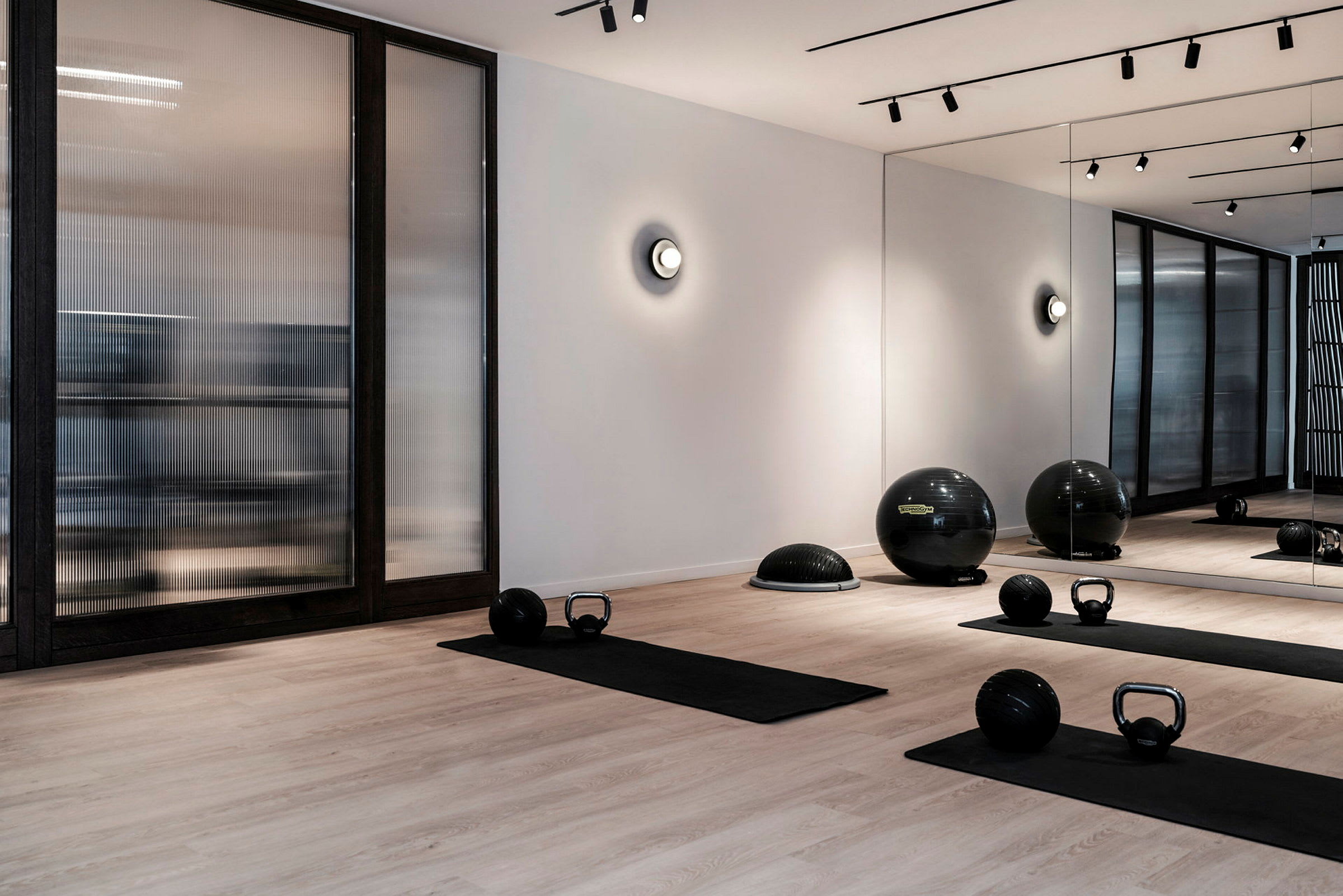 Moderni kuntosali, jossa on joogamattoja, kahvakuulia ja kuntopalloja puulattialla. Seinällä on peilejä ja lasipaneeleja. (Tekoälyn luoma teksti)