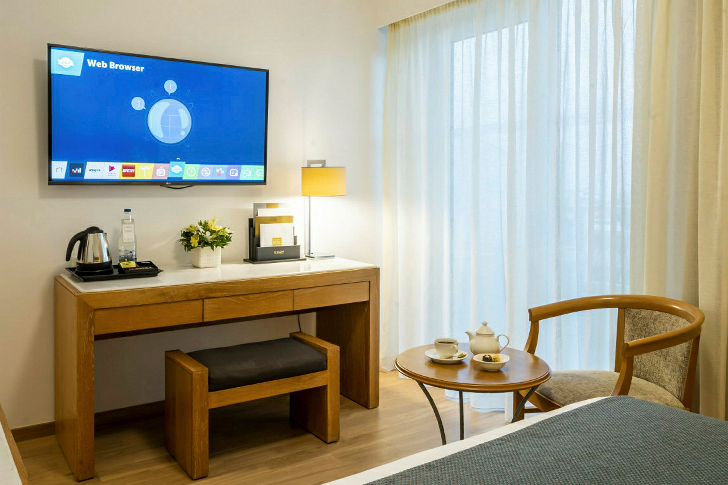 Hotellihuone, jossa on seinälle asennettu televisio työpöydän yläpuolella. Pöydällä on tuoli ja vedenkeitin. Työpöydän vieressä on sivupöytä, jolla on teekuppeja ja tuoli. Taustalla näkyy ikkunoita vaaleiden verhojen takana. (Tekoälyn luoma teksti)