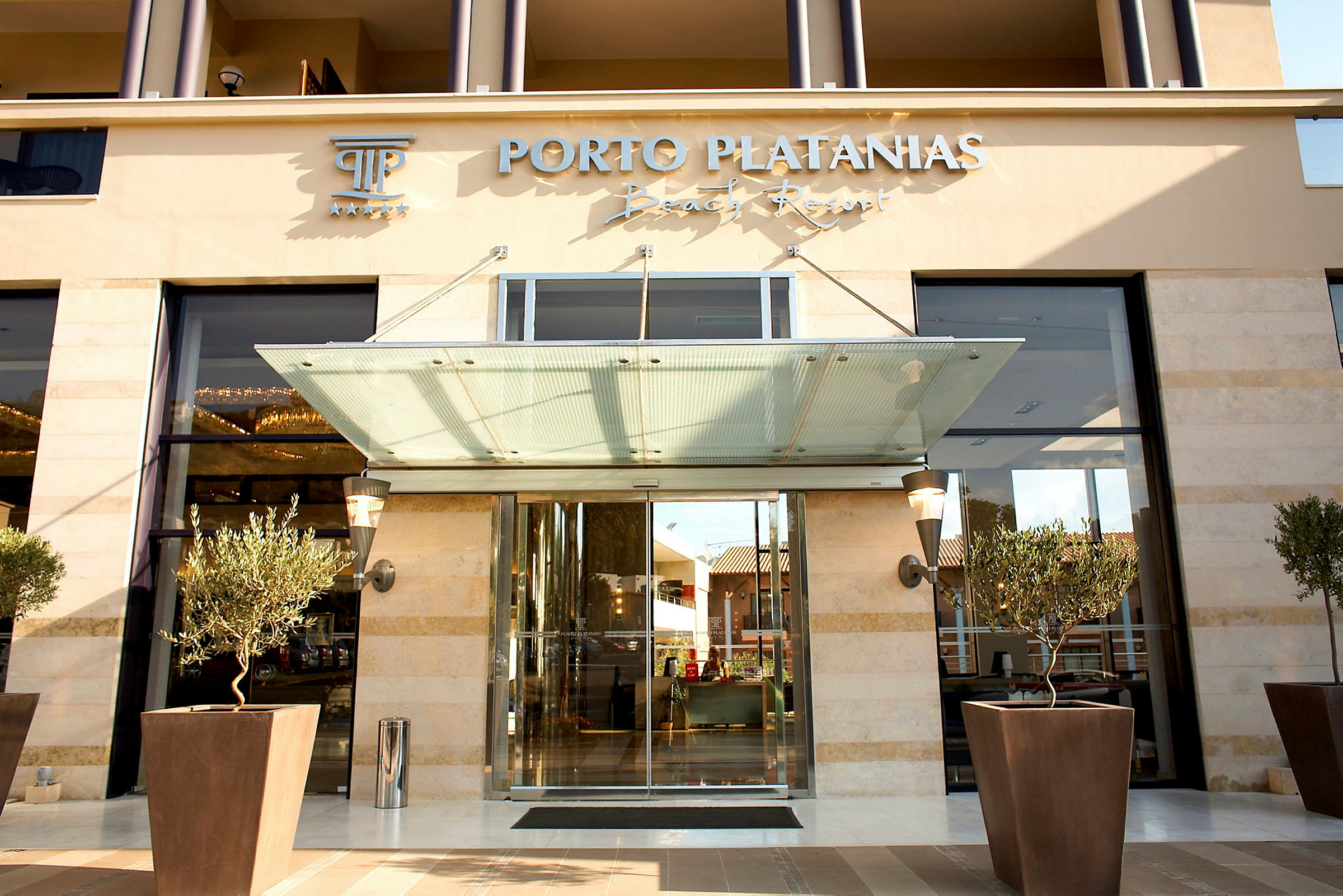 Porto Platanias Beach Resortin sisäänkäynti, lasiovet ja istutetut oliivipuut edessä. (Tekoälyn luoma teksti)