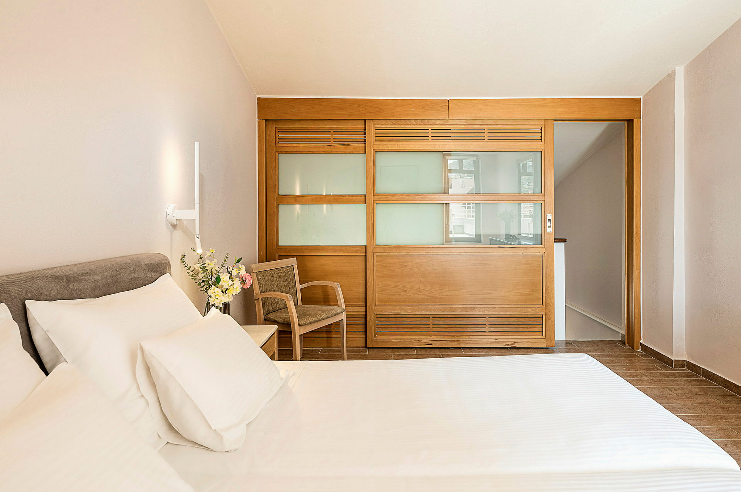 Hotellihuone, jossa on sänky, tuoli ja lasipaneelinen puuseinä. (Tekoälyn luoma teksti)