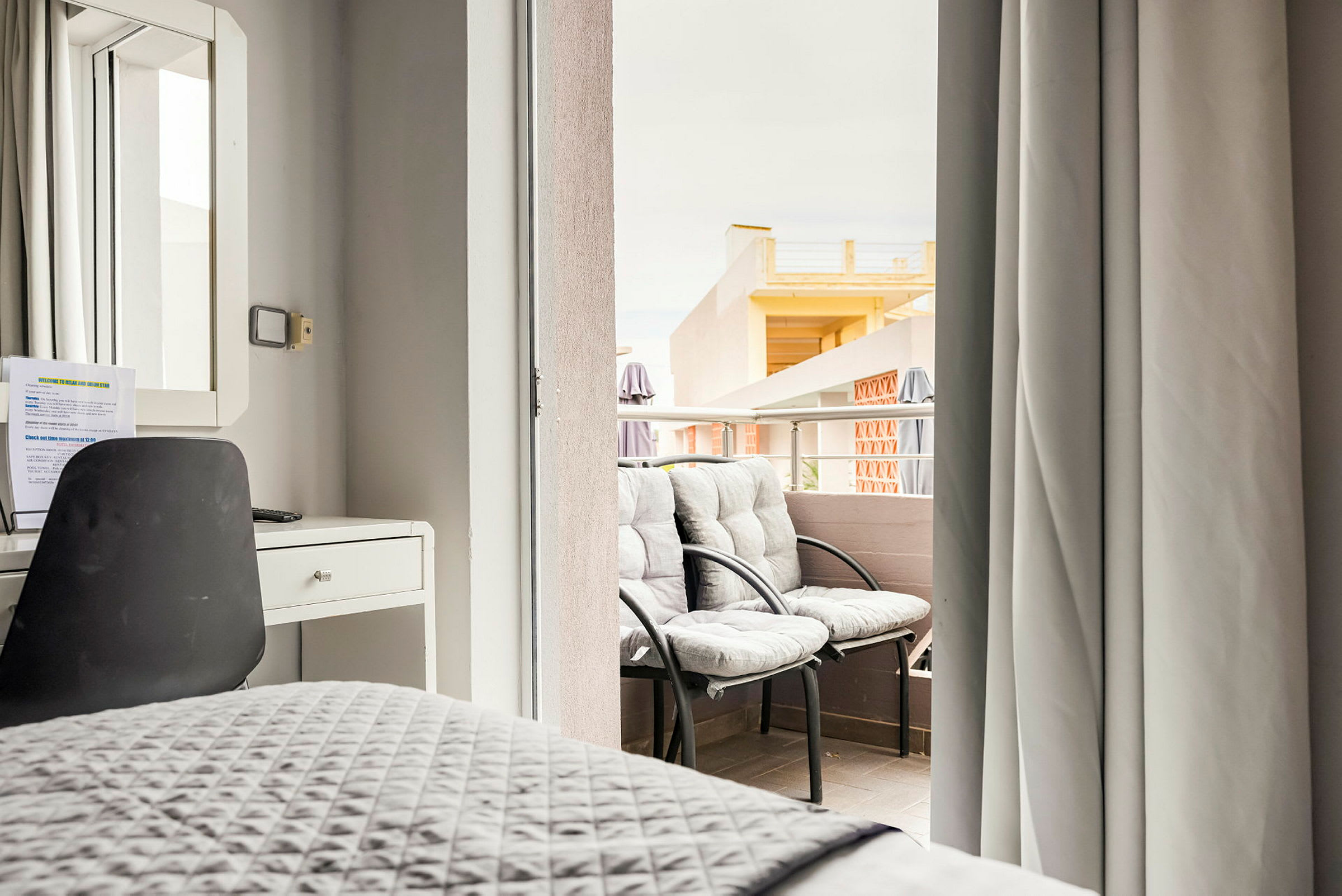 Hotellihuoneen sisustus, jossa työpöytä ja tuoli sekä näkymä parvekkeelle, jolla on kaksi tuolia. (Tekoälyn luoma teksti)