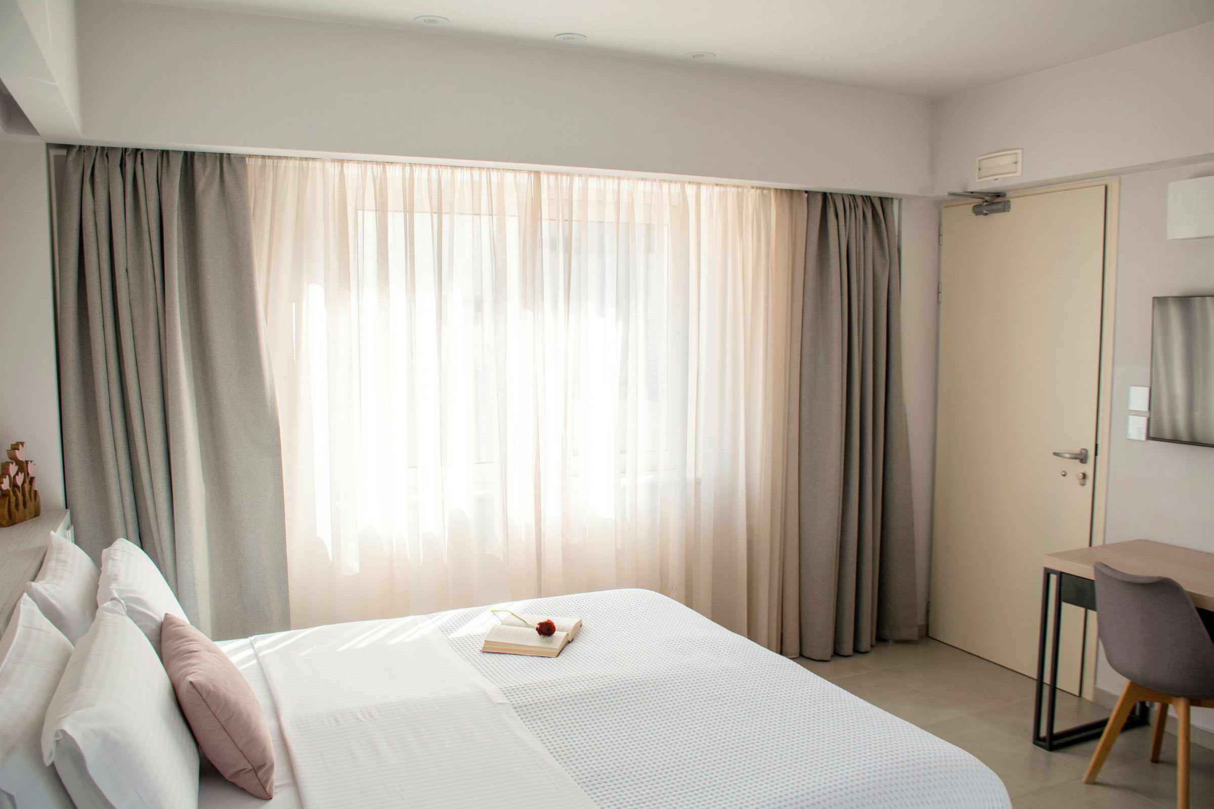 Hotellihuone, jossa on pedattu sänky, ikkuna verhoineen ja kirjoituspöytä tuoleineen. (Tekoälyn luoma teksti)