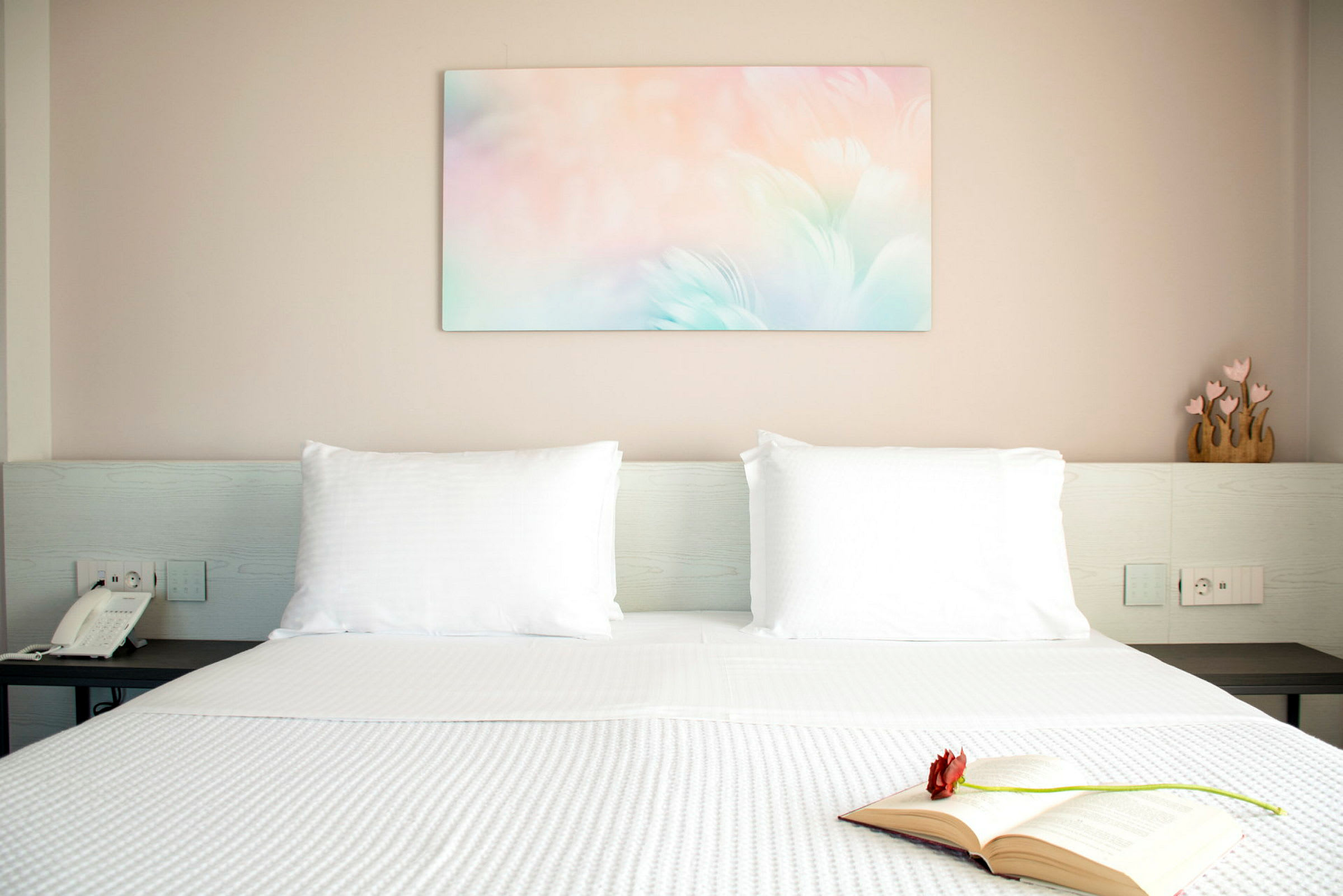 Moderni hotellihuone, jossa on vastapetattu sänky, avoin kirja kukalla ja koristeellinen taulu seinällä. (Tekoälyn luoma teksti)