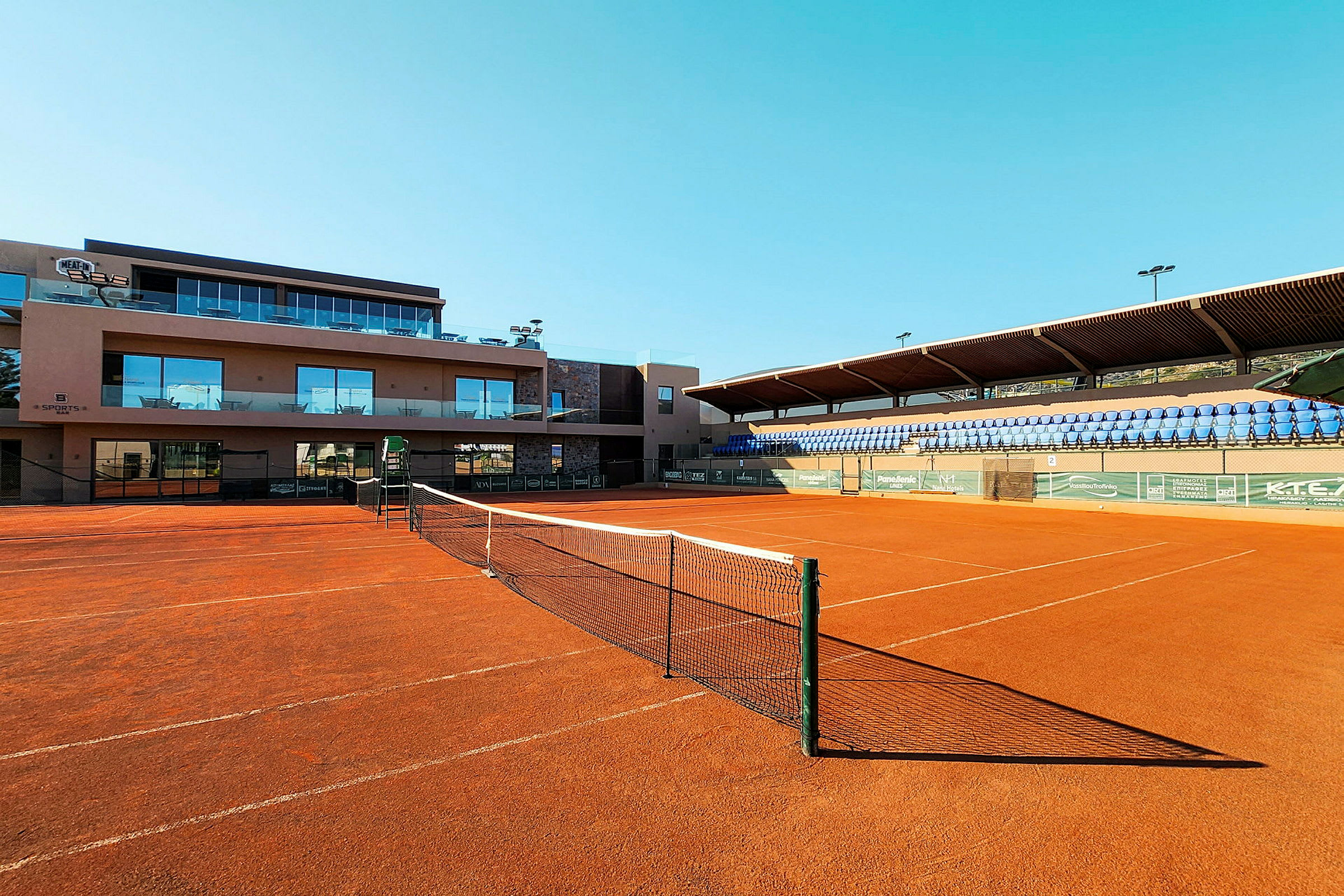 Tenniskenttä punaisella soralla ja verkolla rakennuksen ja katsomoiden edessä. (Tekoälyn luoma teksti)