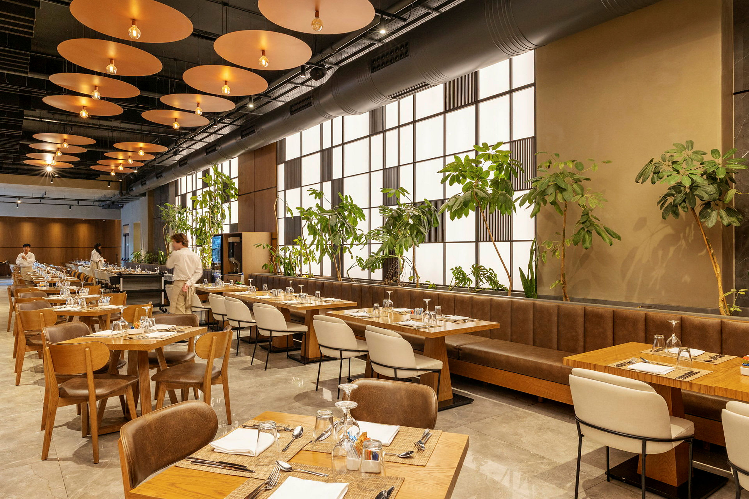 Moderni ravintola, jossa on puupöytiä ja tuoleja, vihreitä kasveja seinän vieressä ja tarjoilija näkyvissä taustalla. (Tekoälyn luoma teksti)