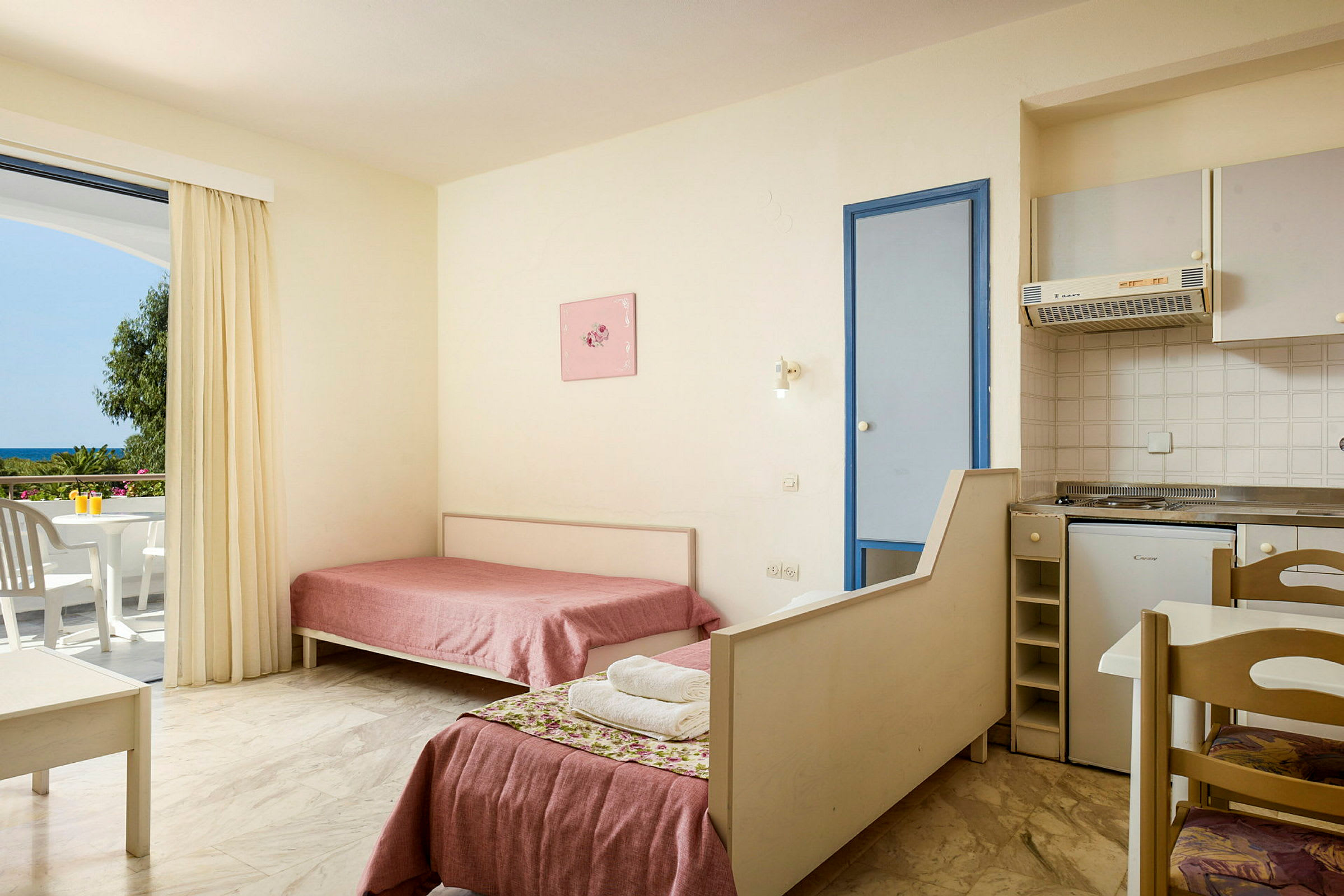 Hotellihuoneen sisätila, jossa kaksi yhden hengen vuodetta, pieni keittiö ja parveke näkymällä. (Tekoälyn luoma teksti)