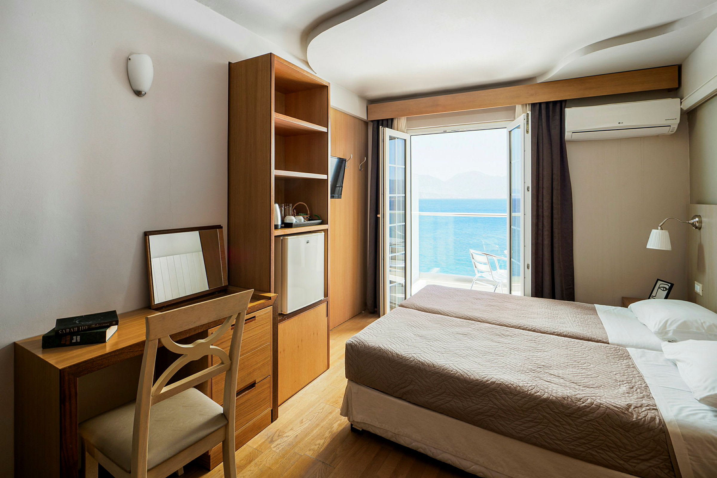 Hotellihuone, jossa kaksi yhden hengen vuodetta, kirjoituspöytä ja tuoli sekä kaappi, jossa on jääkaappi. Ulkona näkyy parveke merinäköalalla. (Tekoälyn luoma teksti)