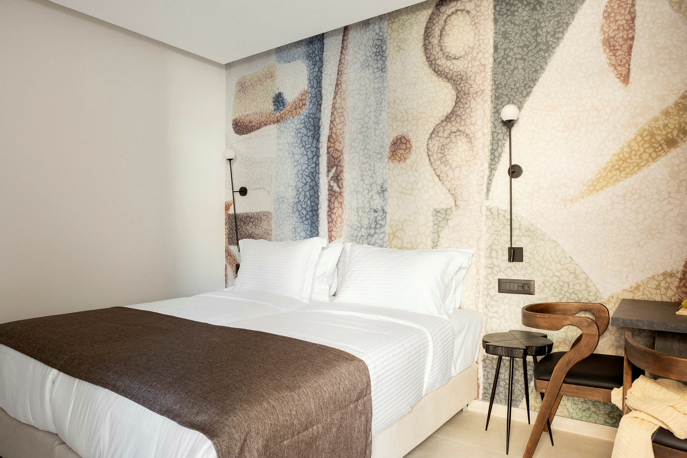 Hotellihuone, jossa kaksi sänkyä valkoisilla vuodevaatteilla ja ruskealla peitteellä. Sängyn pääty on koristeltu värikkäällä taustalla. (Tekoälyn luoma teksti)