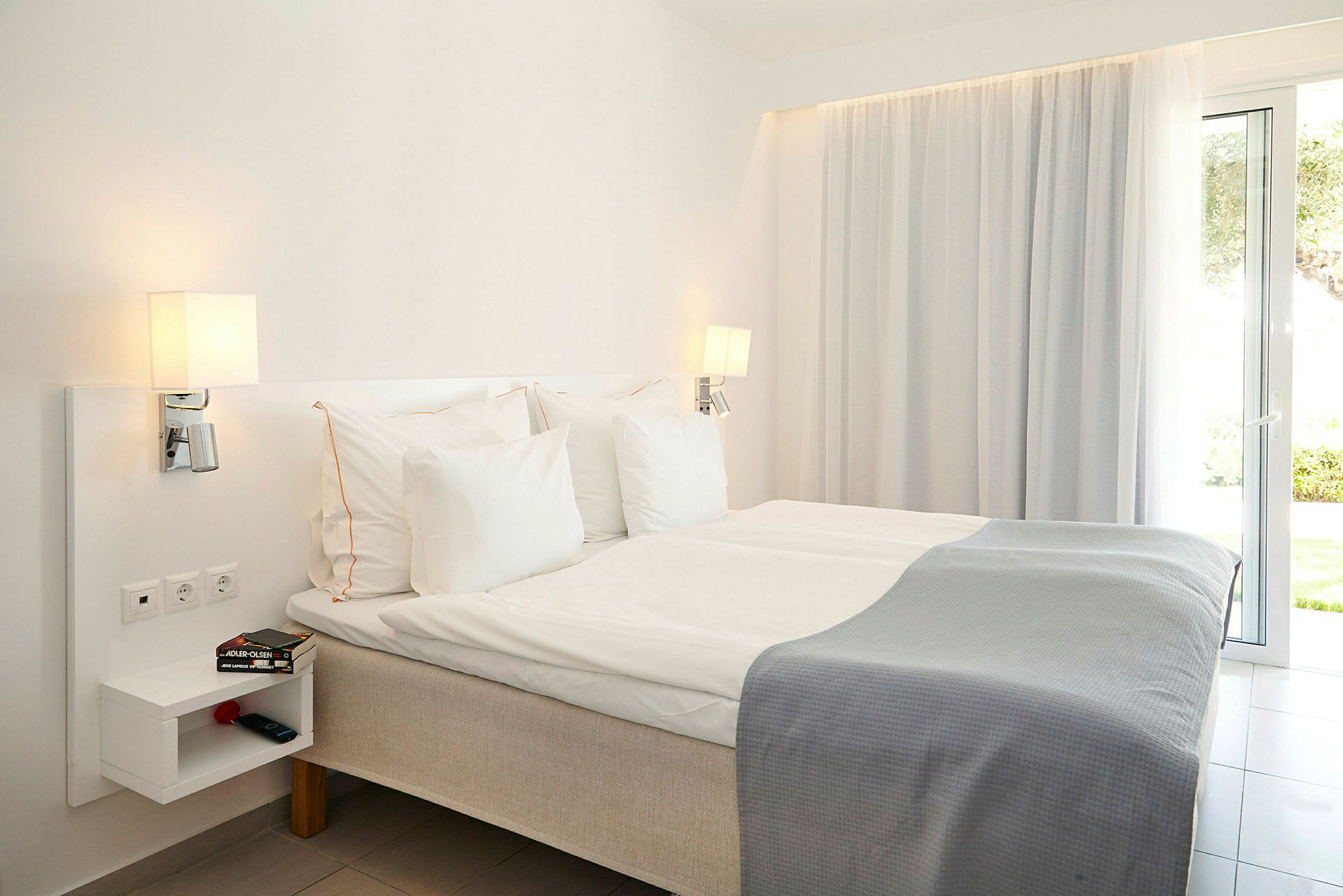 Hotellihuone, jossa sänky, valkoiset vuodevaatteet ja harmaa viltti, lamput, hylly ja ikkuna. (Tekoälyn luoma teksti)