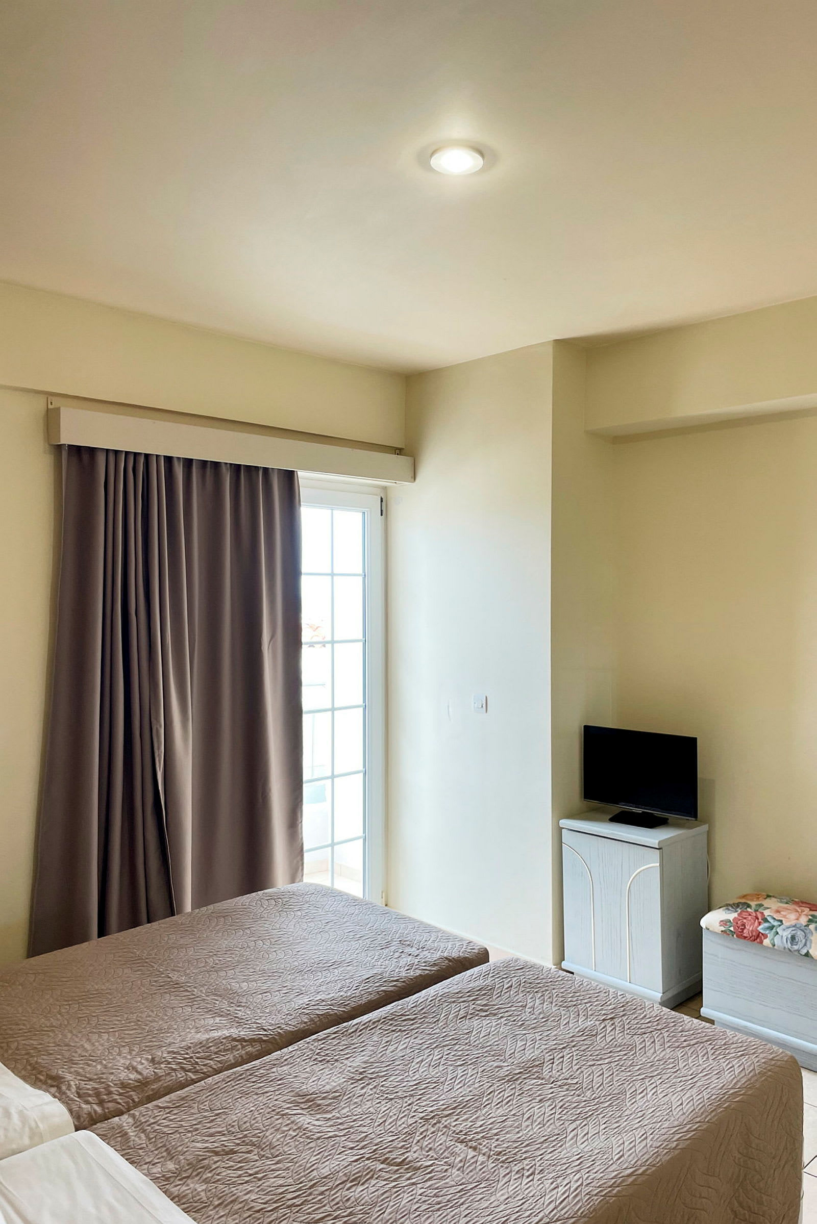 Hotellihuone, jossa kaksi erillisvuodetta, parvekkeen ovi verhoilla ja pieni televisio kaapin päällä. (Tekoälyn luoma teksti)