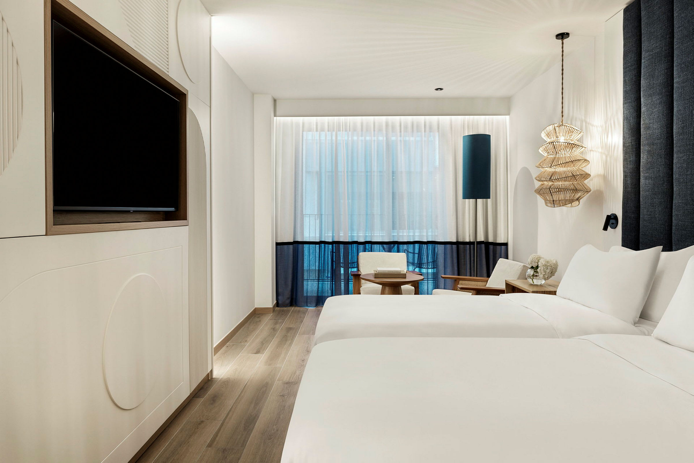 Moderni hotellihuone, jossa on kaksi yhden hengen vuodetta, seinällä televisio, istuimet ja koristeellinen lamppu. (Tekoälyn luoma teksti)