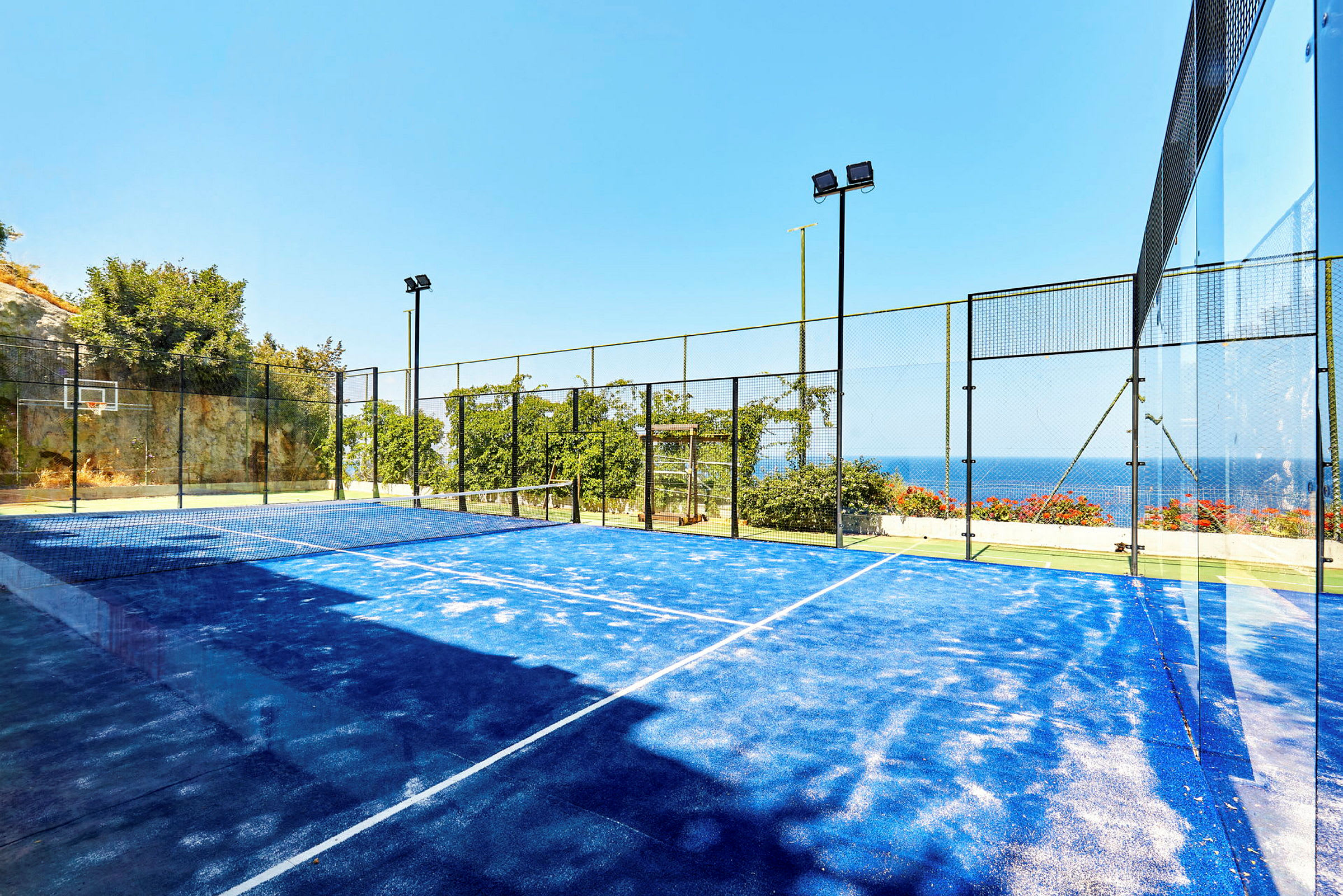 Padelkenttä, jossa on sininen alusta, ympäröity aidalla ja näkymä merelle. (Tekoälyn luoma teksti)