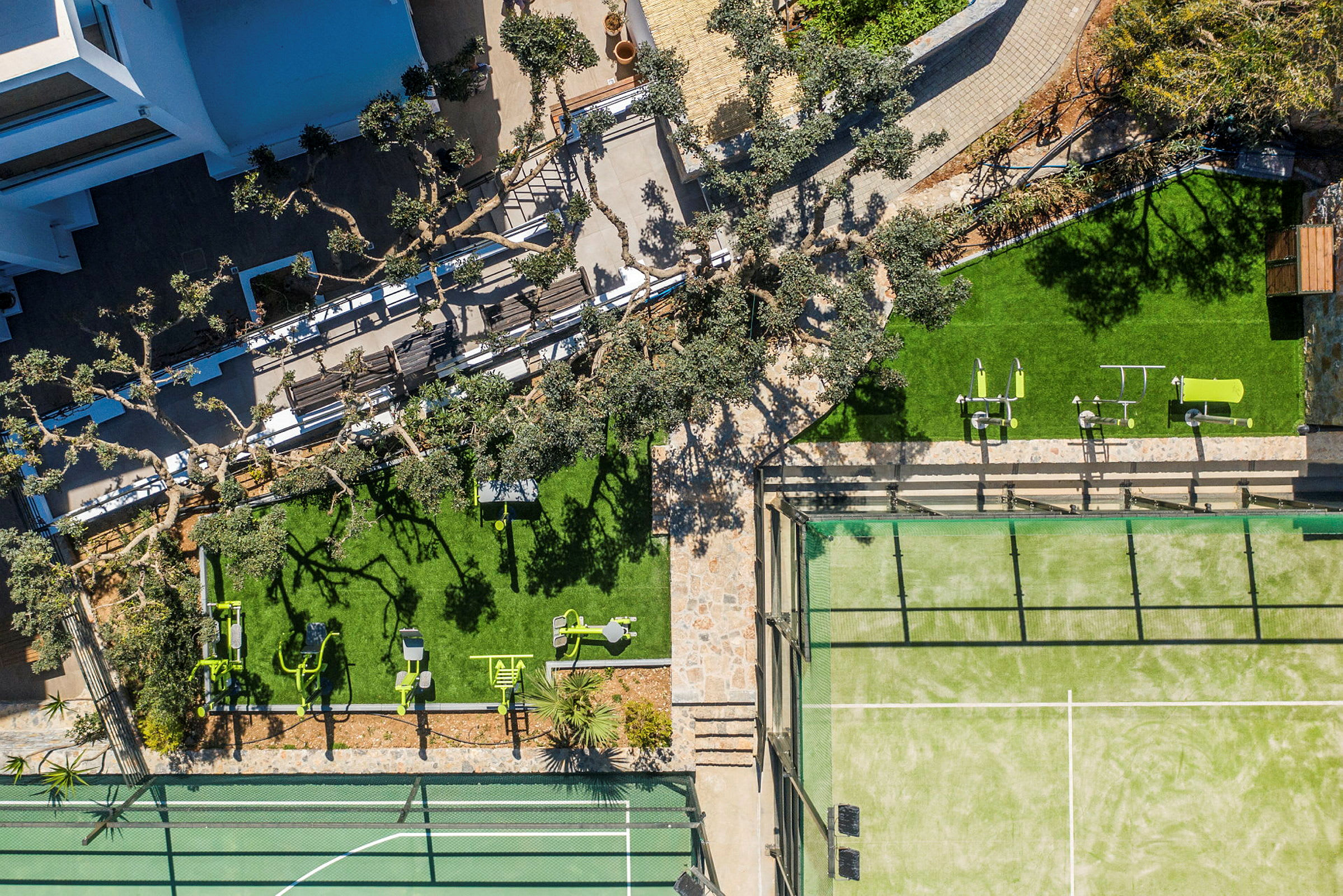 Ilmakuva ulkoalueesta, jossa on puita, vihreitä nurmikoita, kuntoiluvälineitä ja tennisrata. (Tekoälyn luoma teksti)