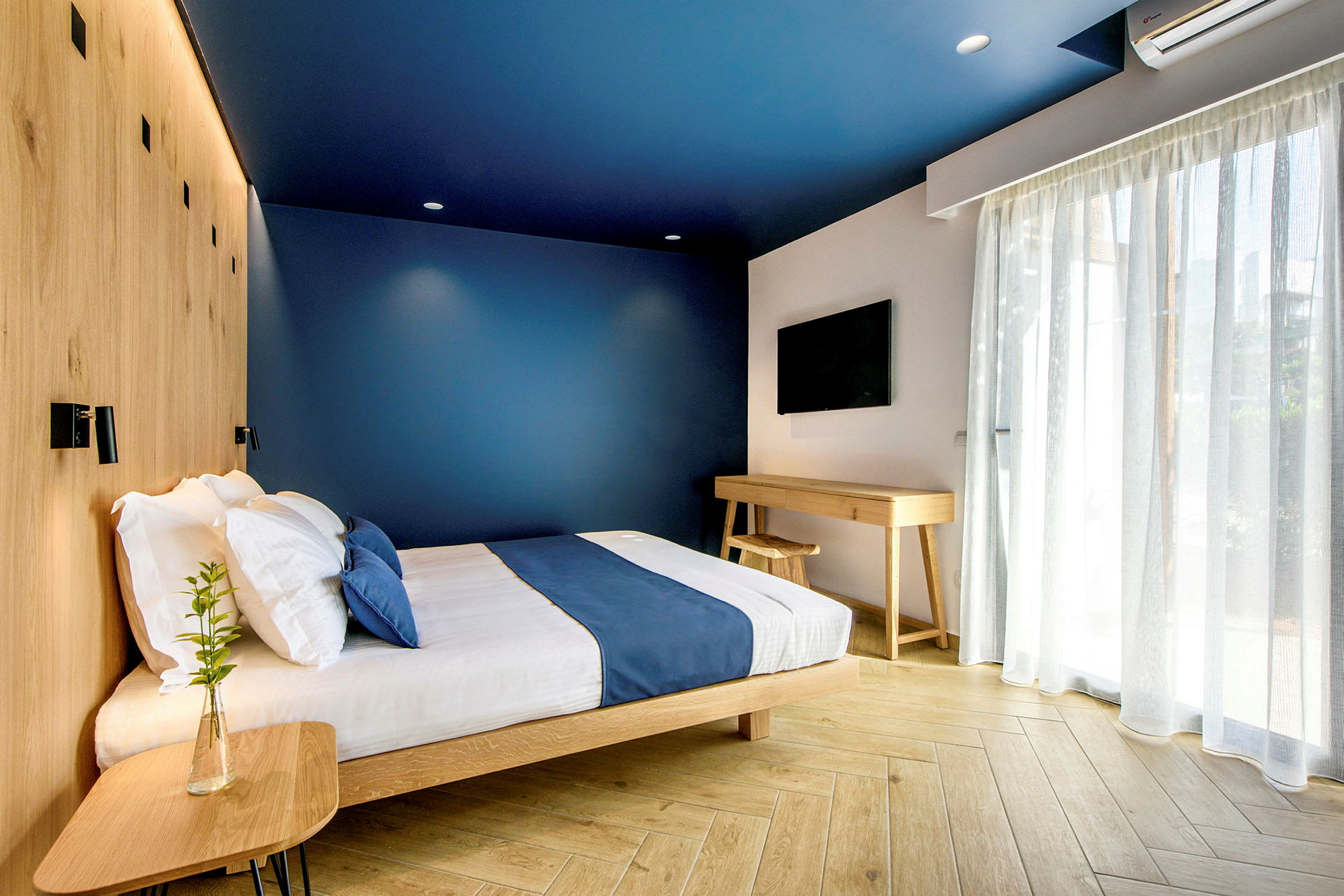 Moderni hotellihuone, jossa siniset seinät, sänky sinisillä vuodevaatteilla ja puukalusteet. (Tekoälyn luoma teksti)