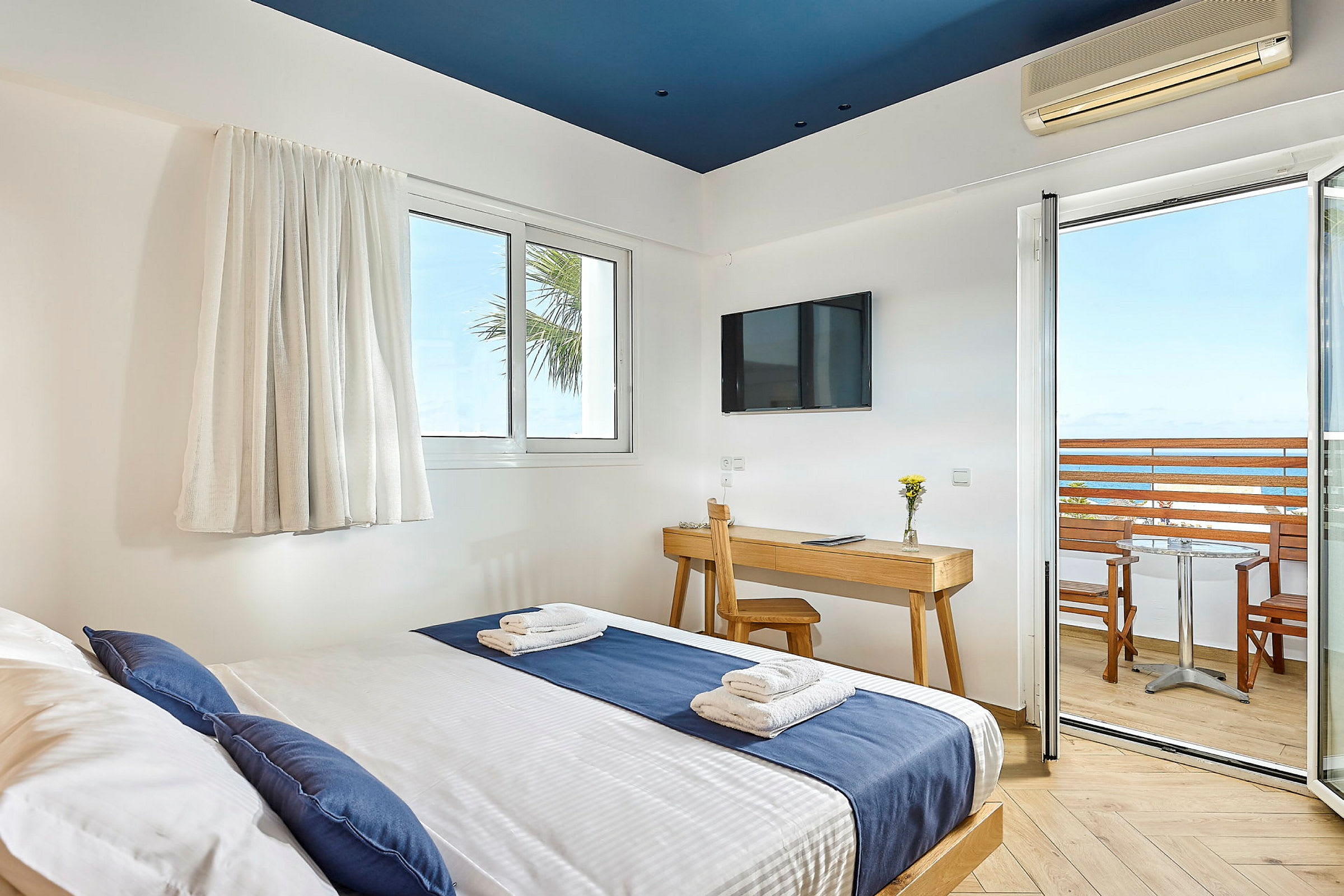 Hotellihuone, jossa sänky sinisen ja valkoisen vuodevaatteet, kirjoituspöytä tuolilla, televisio seinällä ja parveke merinäköalalla. (Tekoälyn luoma teksti)