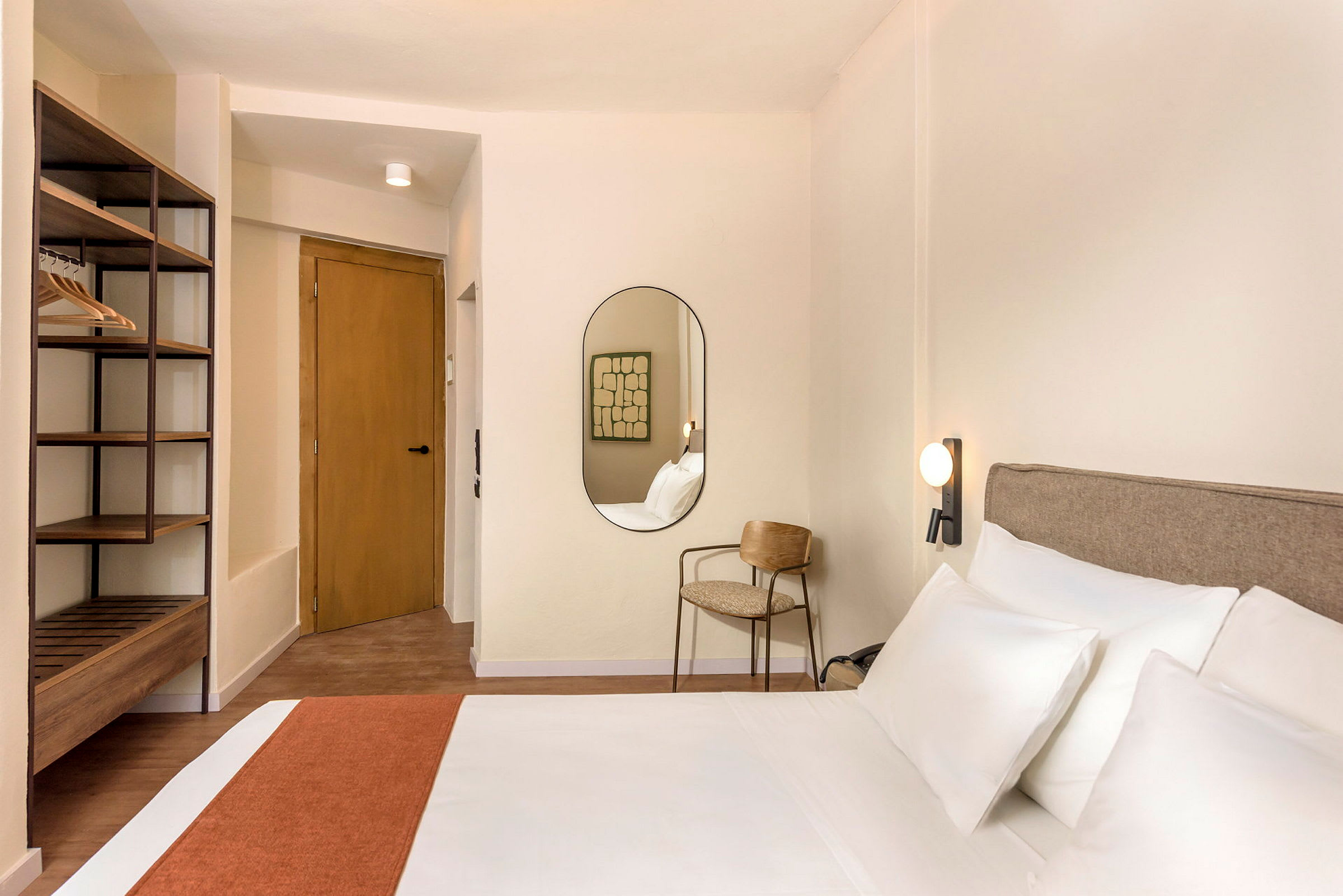 Hotellihuone, jossa on sänky, peili, tuoli ja avonainen vaatesäilytys. (Tekoälyn luoma teksti)