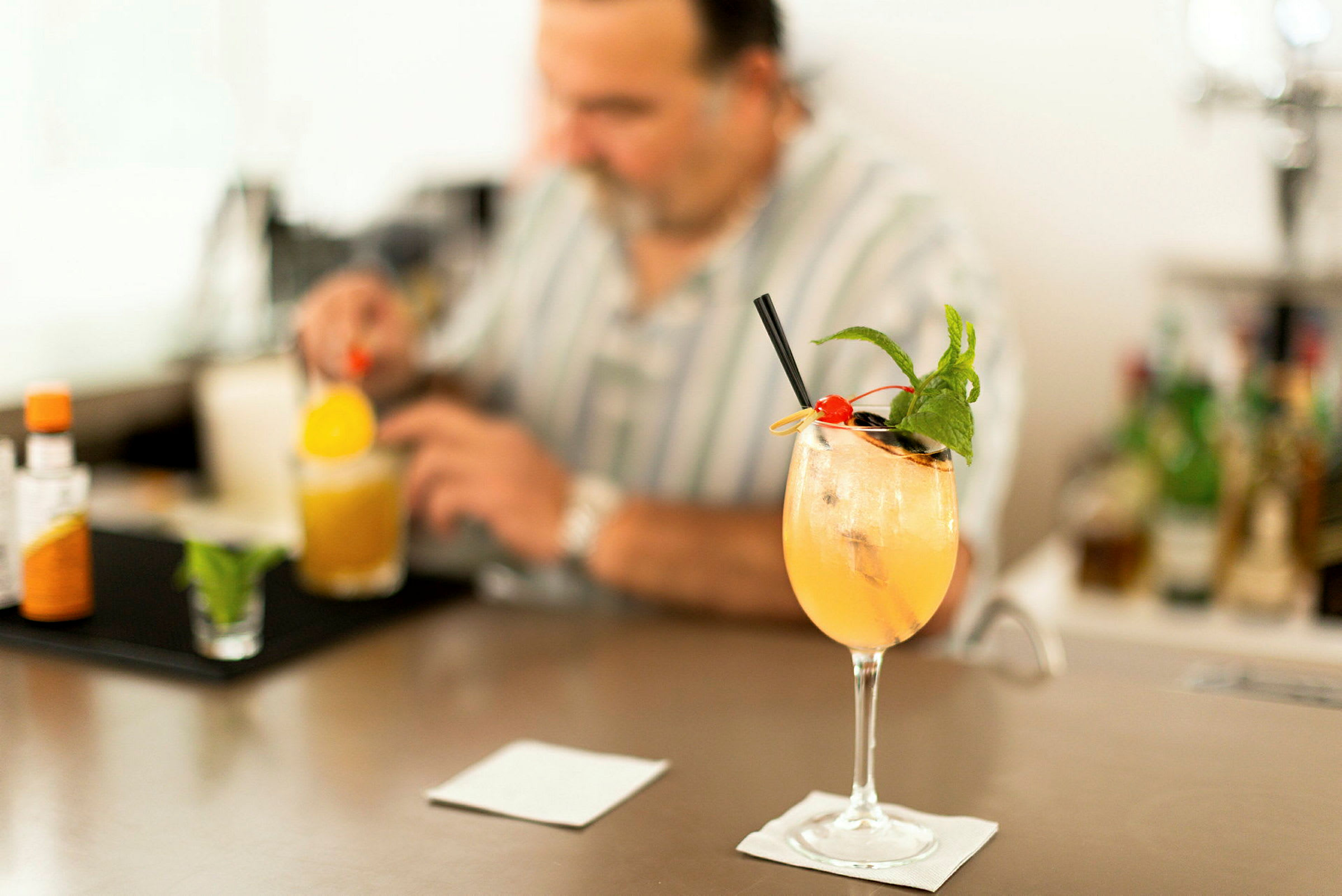 Cocktail-lasi, jossa värikästä juomaa, koristeltu jääpaloilla, mintulla ja kirsikalla, seisoo baaritiskillä. Taustalla henkilö valmistaa juomaa. (Tekoälyn luoma teksti)
